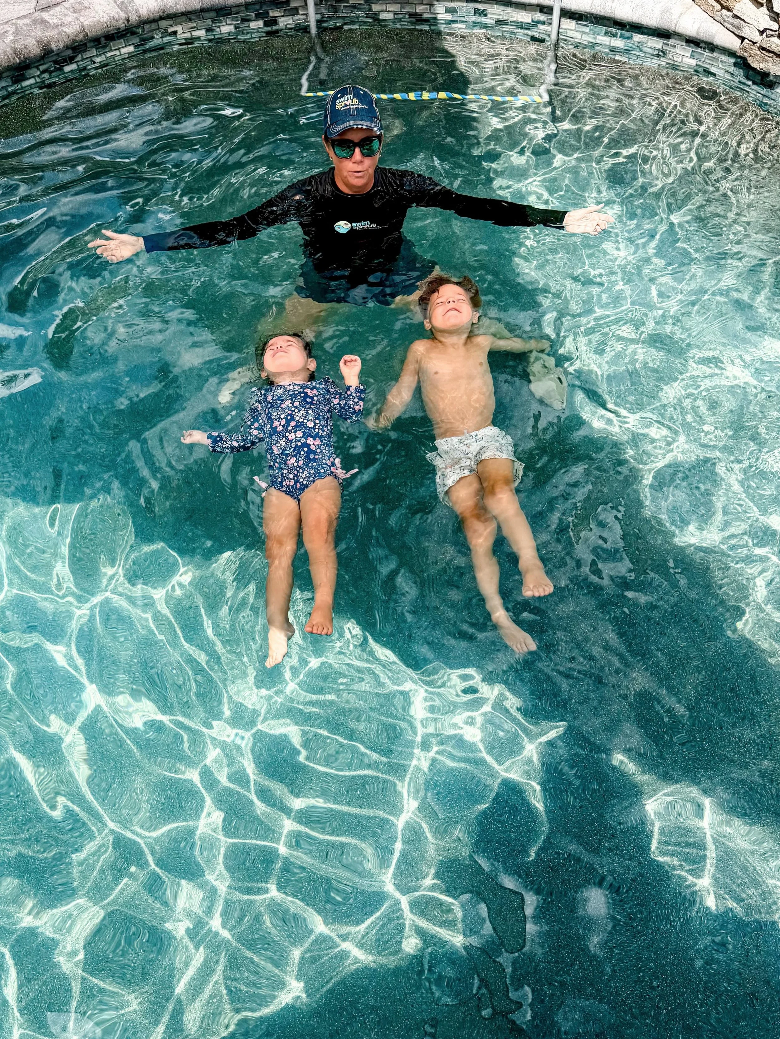 A woman in a black swim shirt and cap is floating in a swimming pool with her arms outstretched. Two children, a girl in a black floral swimsuit and a boy in swim trunks, are lying back in the water with their eyes closed, enjoying the sun.