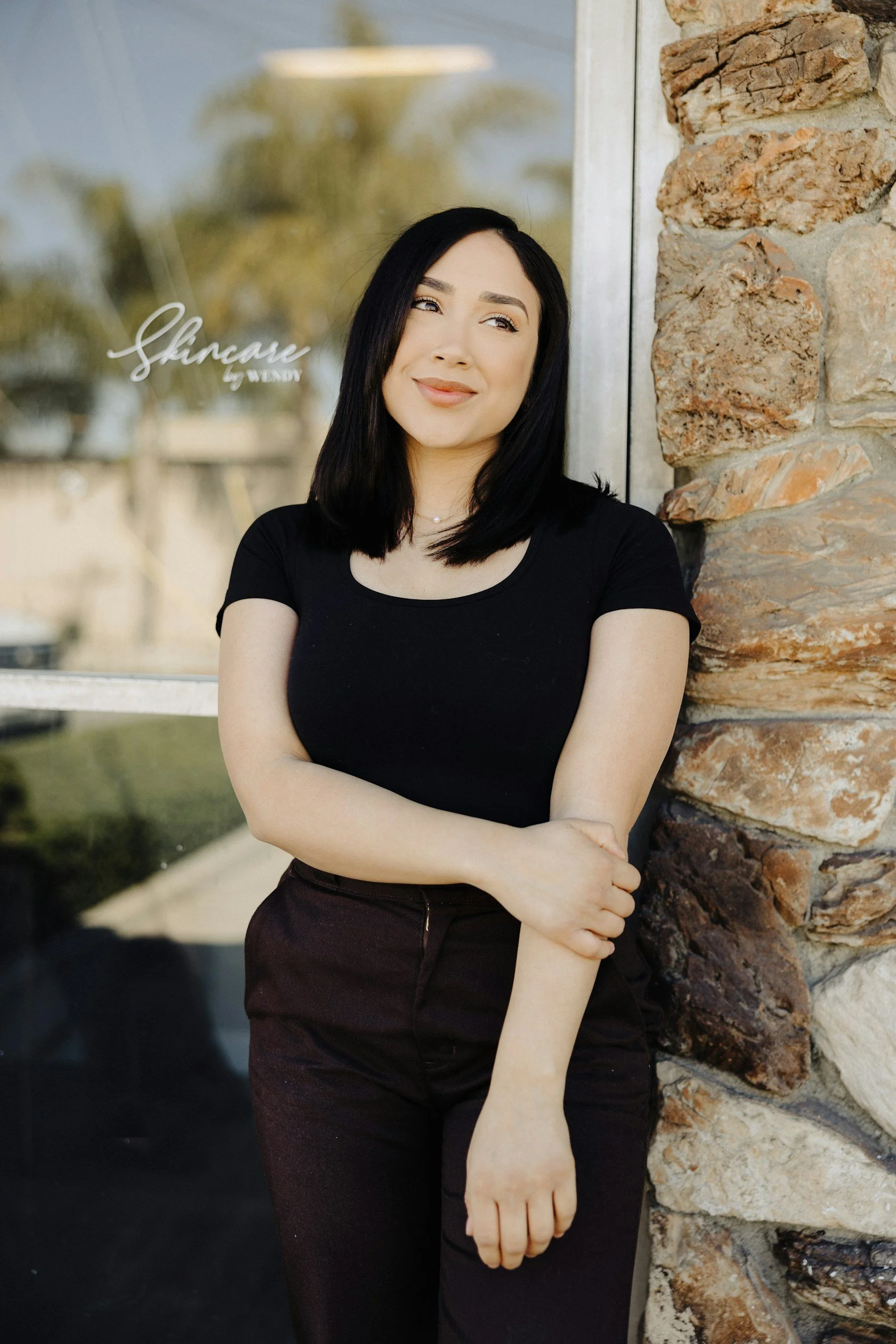 A woman with black hair wearing a black top and dark skirt standing against a stone wall with glass behind her, smiling and looking to her right.