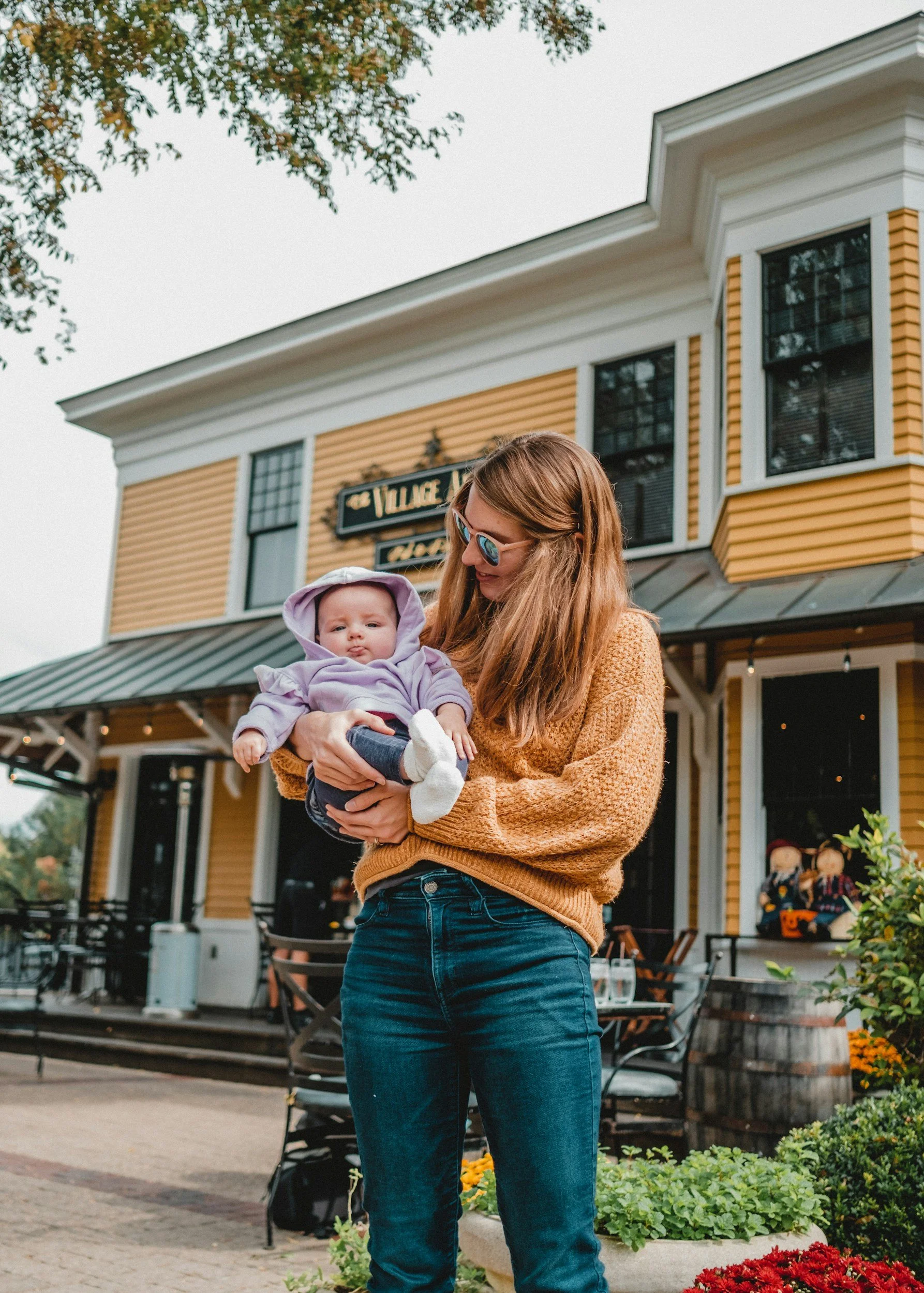Woman with long hair wearing sunglasses holding a baby in a lavender hoodie outside a yellow building with a sign reading "Village" and some outdoor seating.