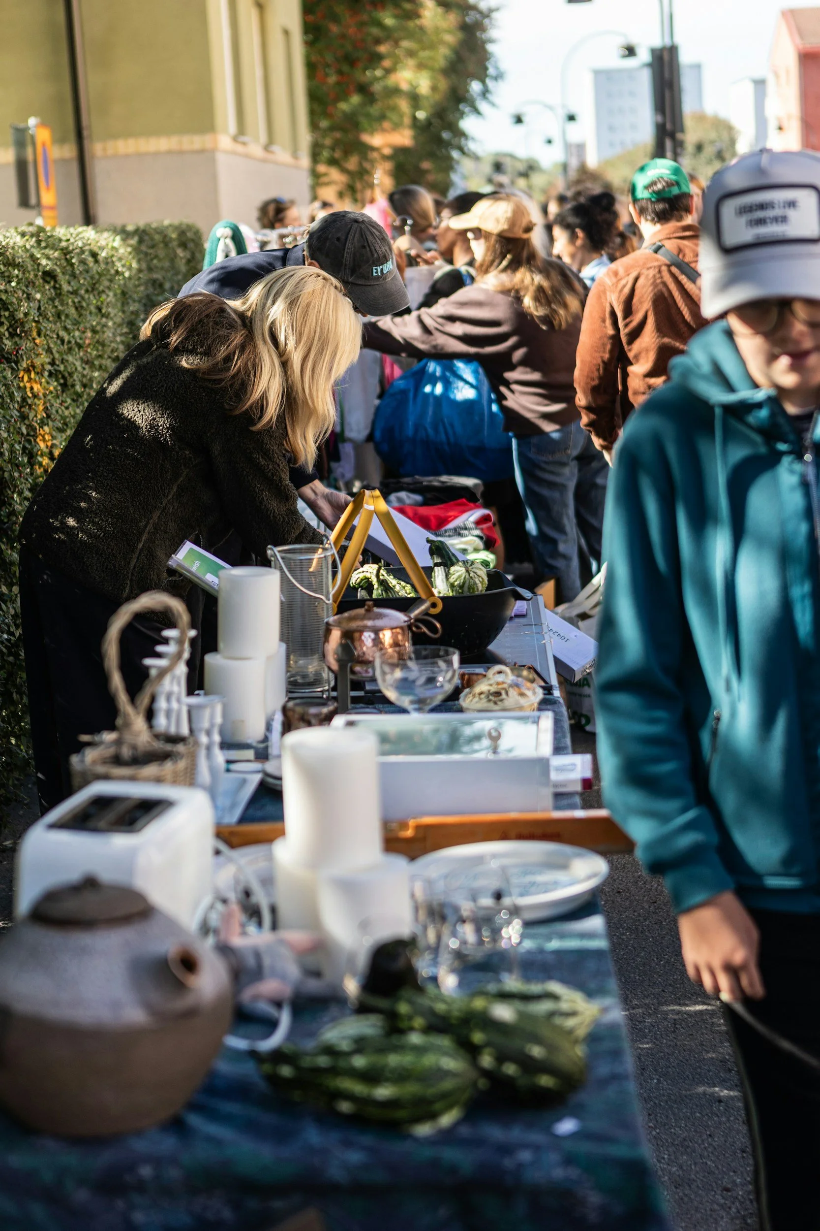 A busy outdoor market scene with people browsing tables filled with various items like candles, ceramics, and decorative objects. The market is set along the street with buildings and trees in the background, and some traders are engaging with customers.
