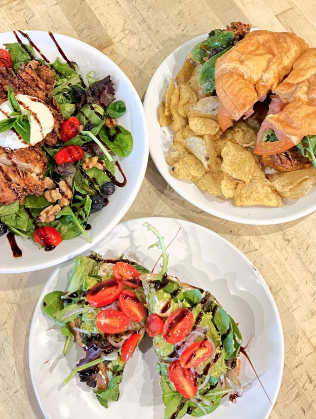 Three dishes on a wooden table: a bowl of potato chips with a sandwich, a salad with berries and nuts, and a salad with cherry tomatoes and greens drizzled with balsamic glaze.