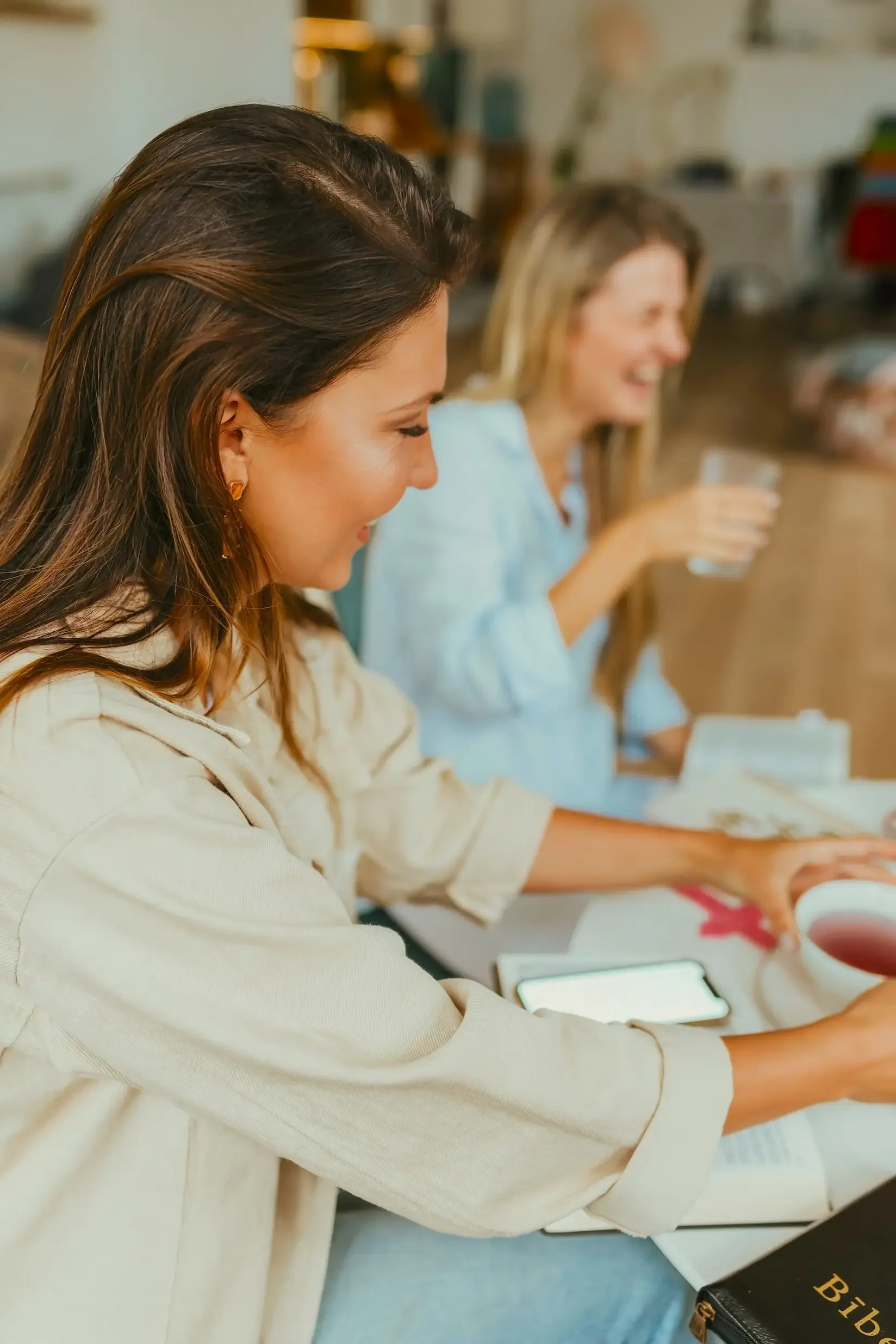 Two women sitting at a table, one has dark hair and the other has blonde hair, both smiling and reaching for items on the table, with cups of tea and a phone visible.