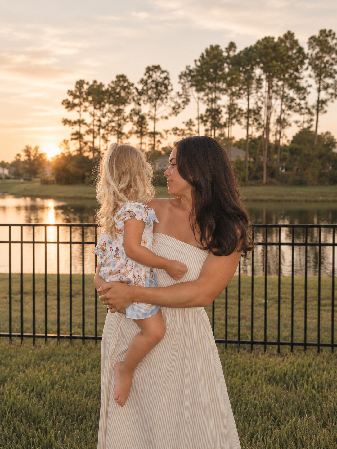 A woman holding a young girl by a lake at sunset, with trees in the background.