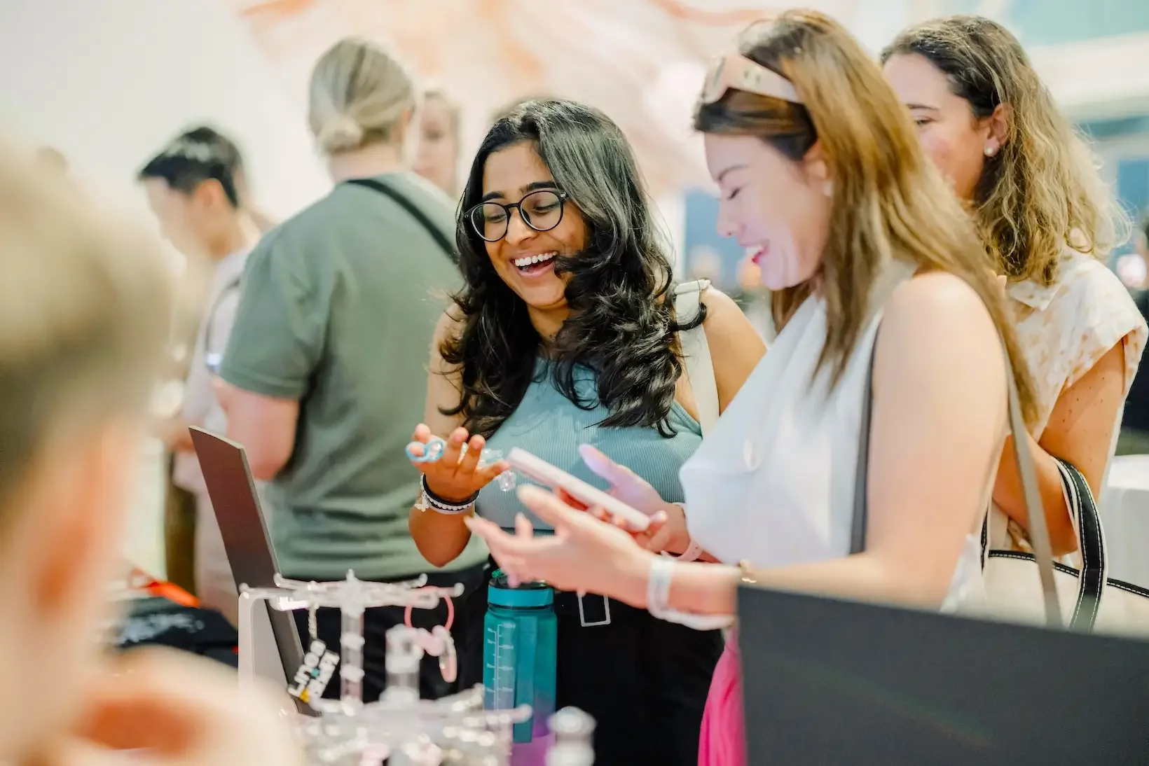 Group of women smiling and talking at an event, with one woman showing her phone to others. Varied women, a display with jewelry or accessories nearby.