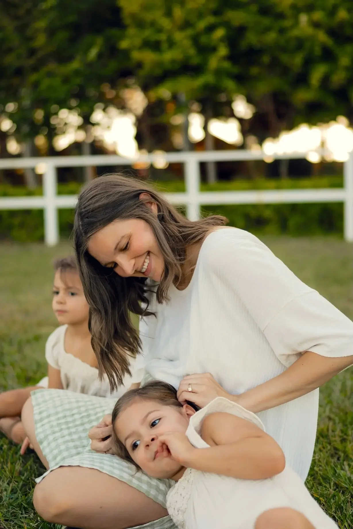 A woman with brown hair smiling with two young girls, one toddler and one slightly older, lying on the grass outdoors near a white fence with trees in the background.