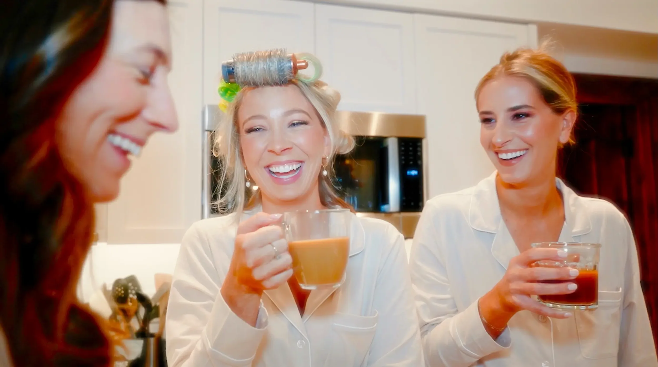 Three women wearing pajamas are smiling and enjoying drinks in a kitchen. One woman, in the center, has hair rollers and is holding a clear glass mug with a hot beverage. The woman on the right is also holding a similar drink. The woman on the left is partially out of frame with long red hair.