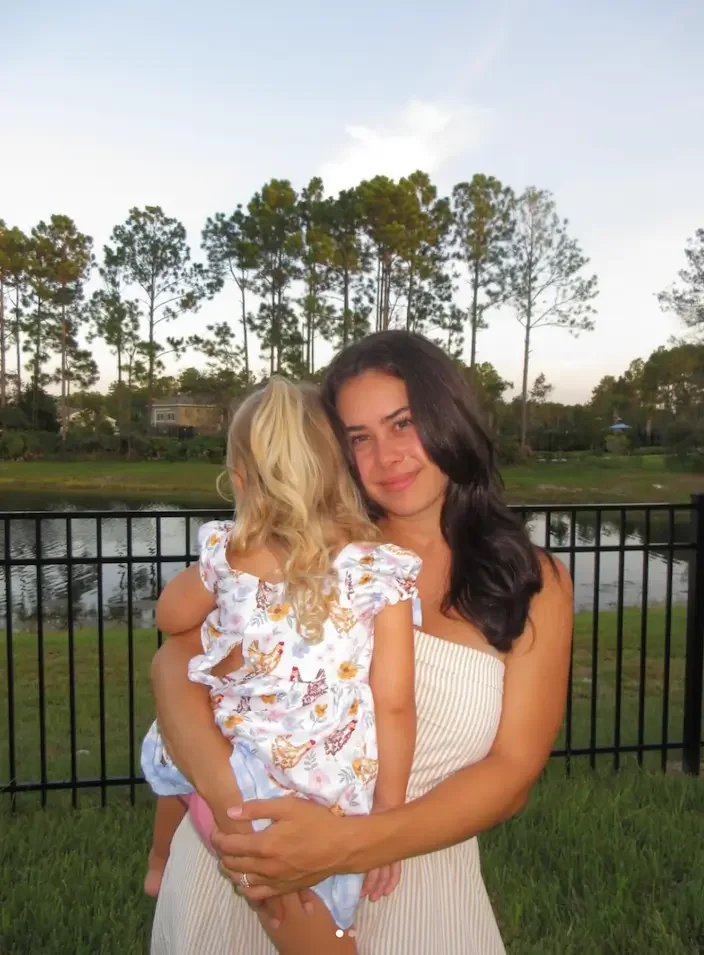 A woman, Mackenzie, holding a young girl near a lake with trees in the background, during late afternoon.