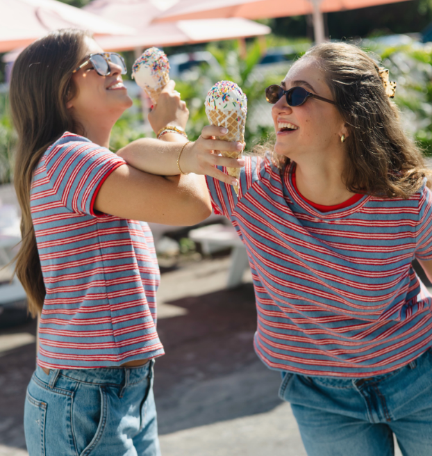 Two women enjoying ice cream cones outdoors on a sunny day, smiling and wearing matching red and blue striped shirts and sunglasses.