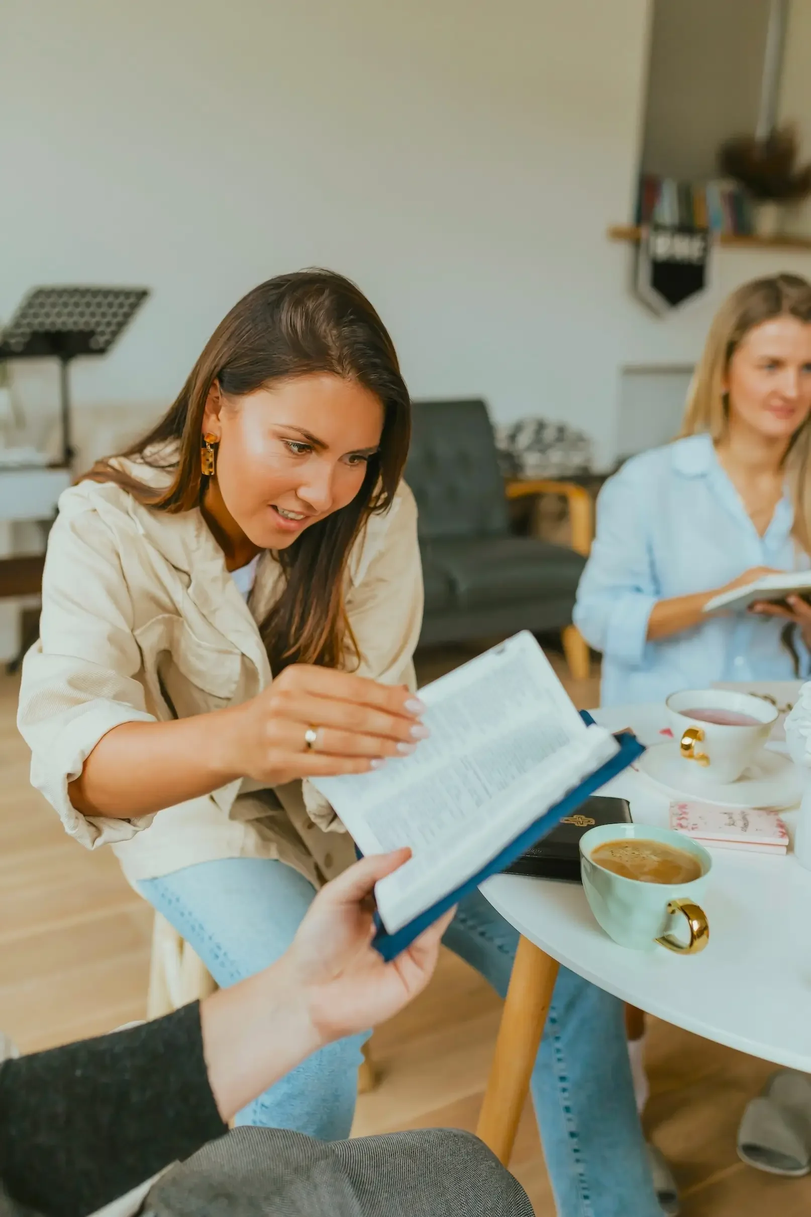 Two women sitting at a table, reading and discussing books, with cups of tea or coffee in front of them, in a cozy indoor setting.