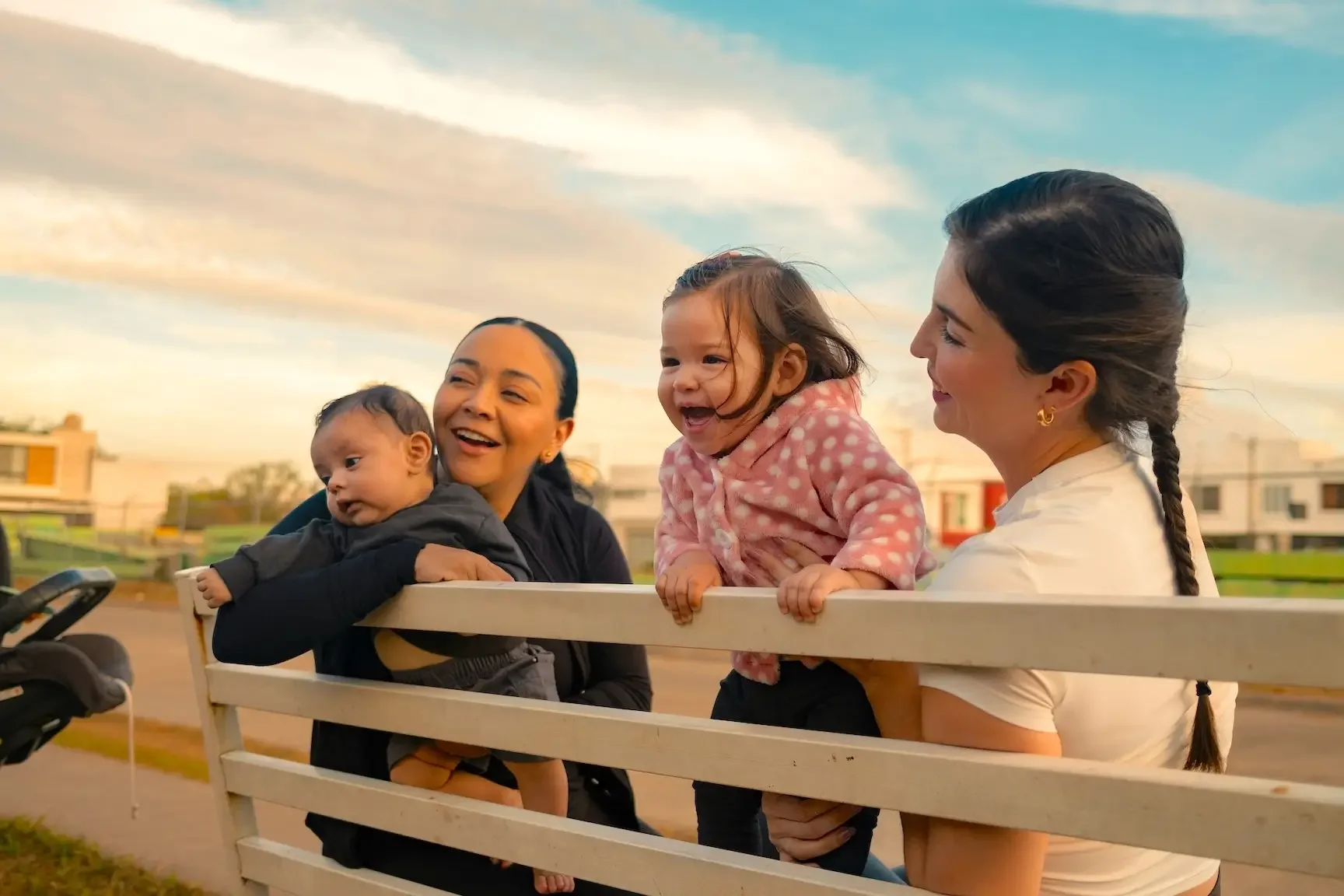 Four people, including two women and two children, smile and laugh on a wooden railing outdoors during sunset. There are houses and a grassy field in the background.