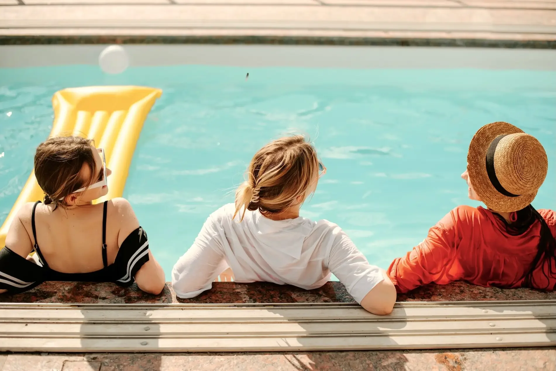 Three women sitting by a swimming pool with a yellow float in the background. One woman is wearing a black bathing suit with a shoulder strap off, another in a white shirt, and the third in an orange jacket and a straw hat, all facing away from the camera.