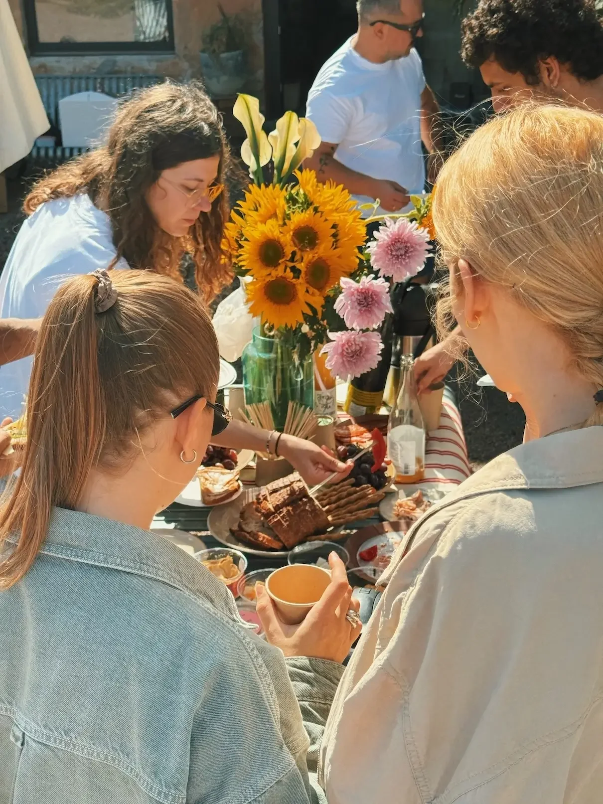 Group of people gathered around a table with food, flowers, and beverages outdoors.