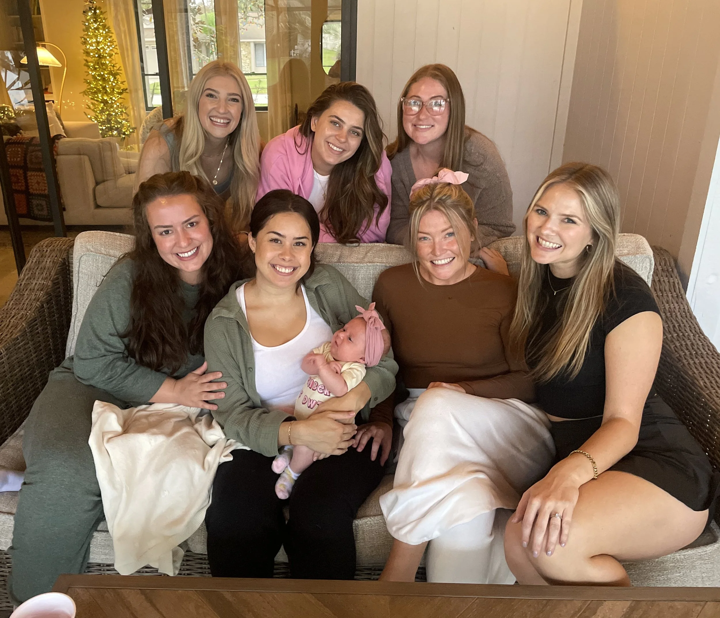 A group of women and a baby sitting and standing together inside a house, smiling at the camera, with a decorated Christmas tree in the background.