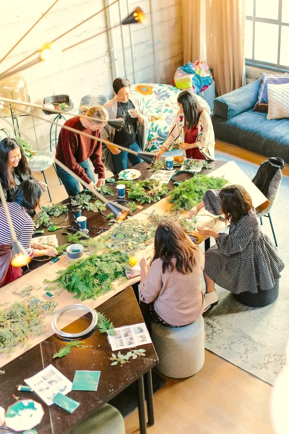 People sitting around a table making flower arrangements with pine branches, leaves, and other natural materials in a cozy cafe space