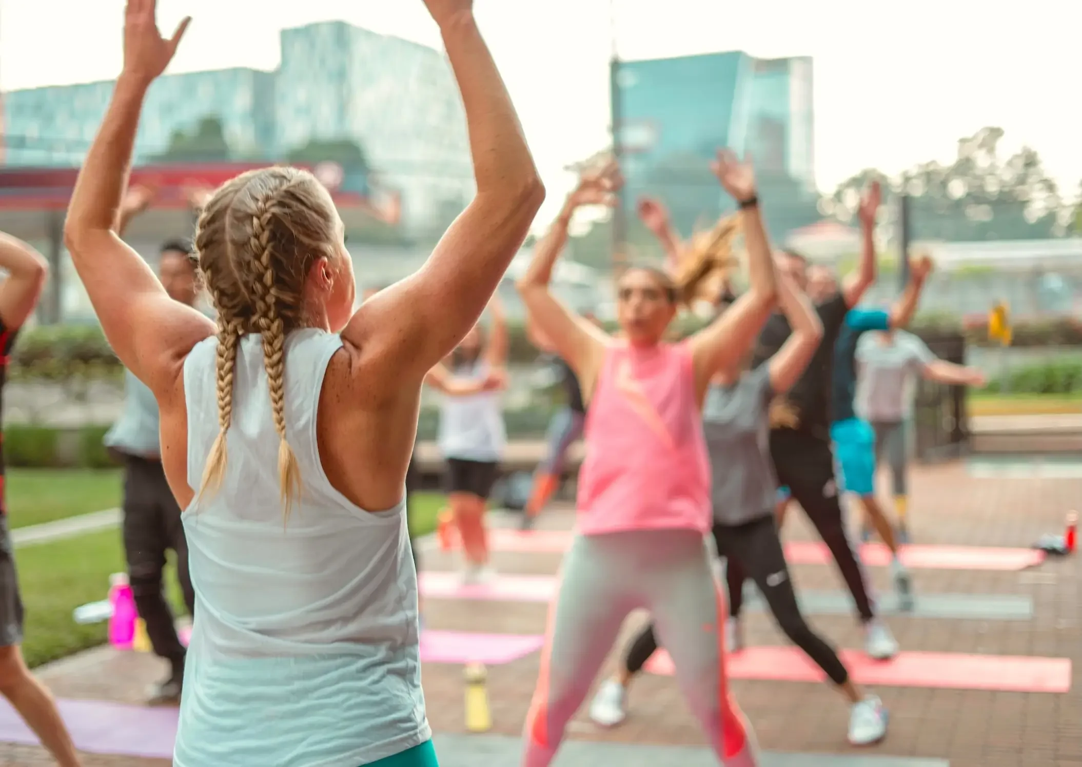 A group exercise class outdoors with women in athletic clothing, arms raised, participating in a workout session.