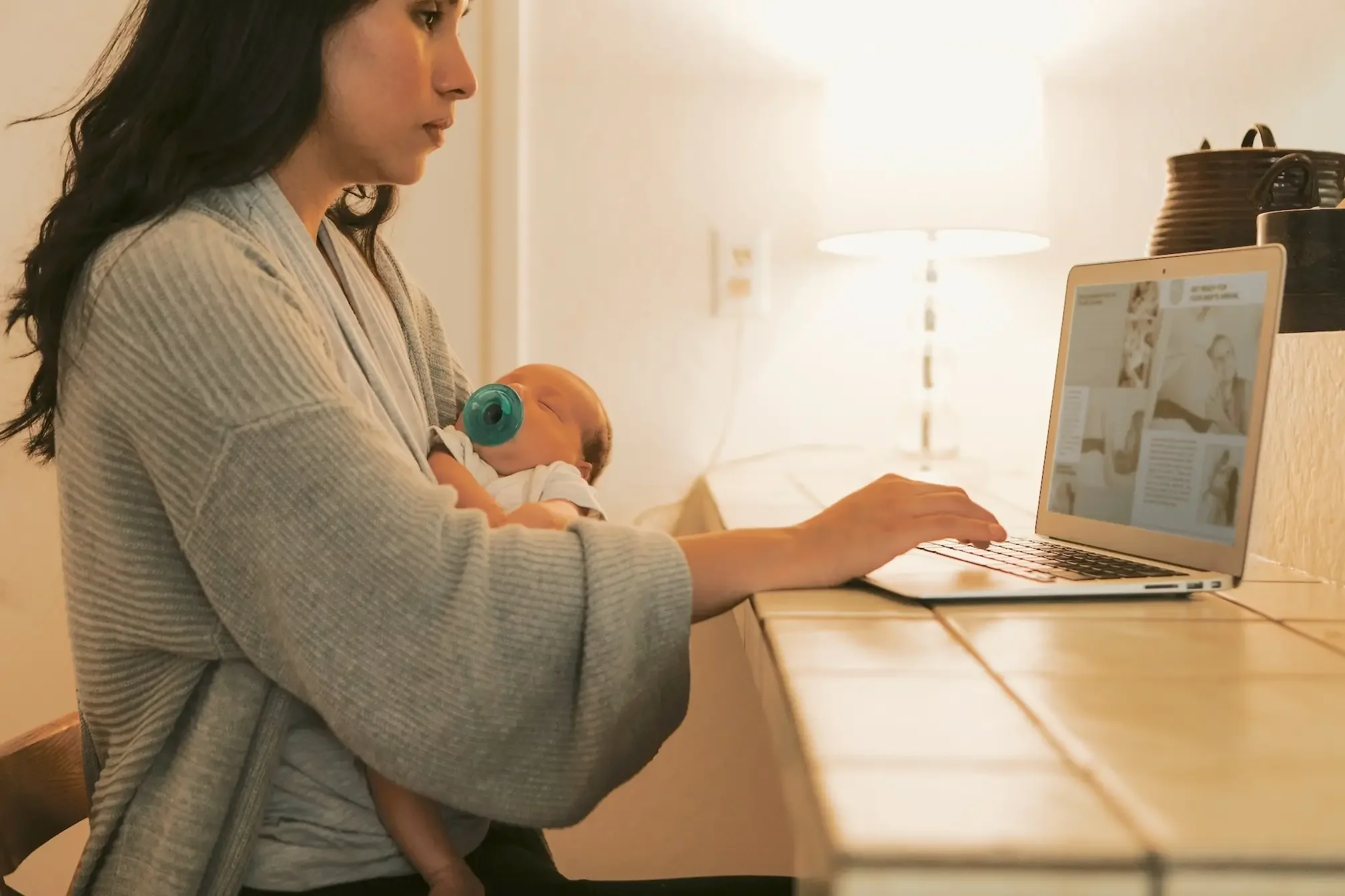 Woman holding a sleeping baby while working on a laptop at a wooden table in a cozy, softly lit room.