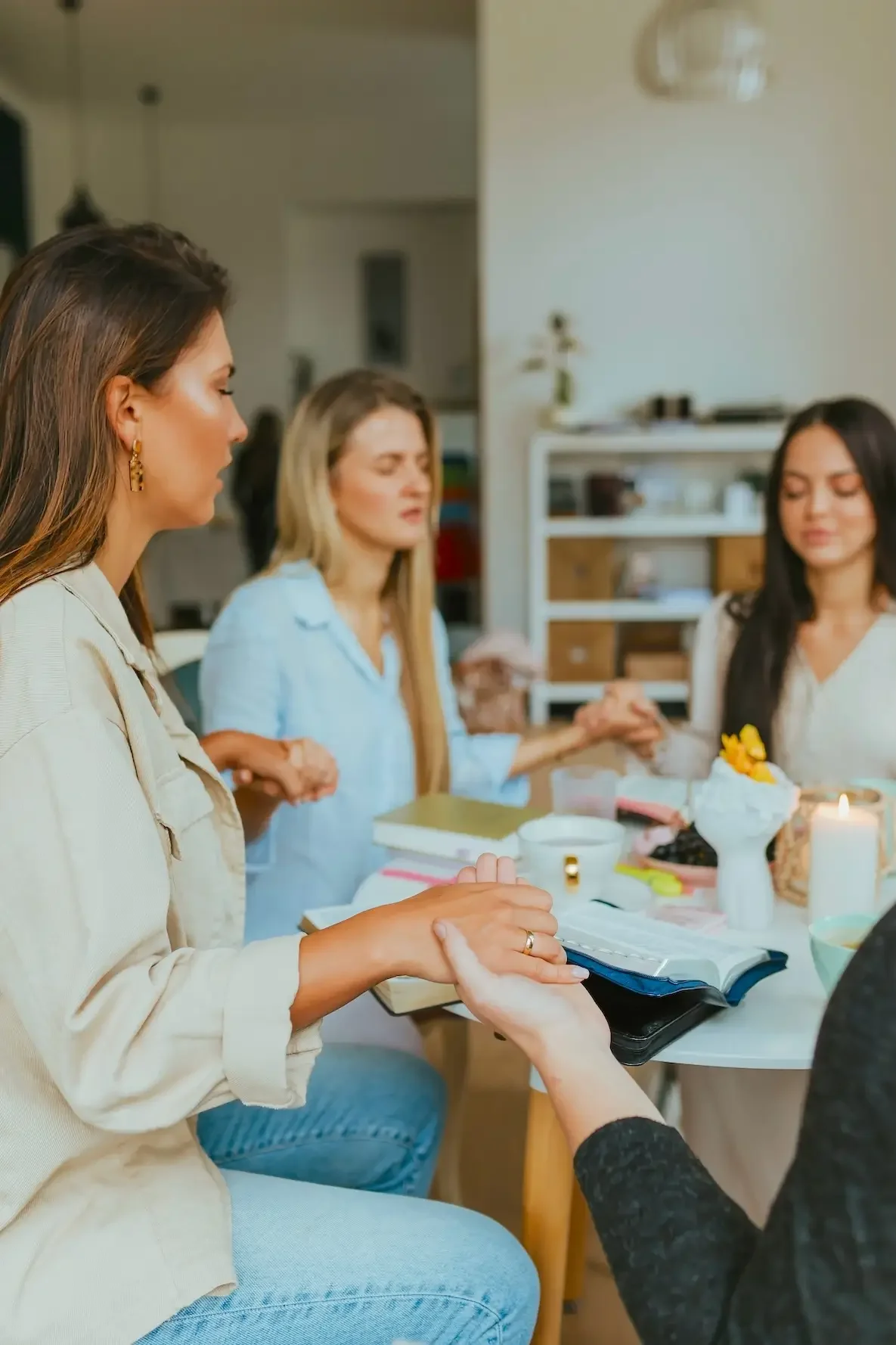 A group of women sitting at a table during a gathering, with some holding hands and a person exchanging a ring or jewelry.