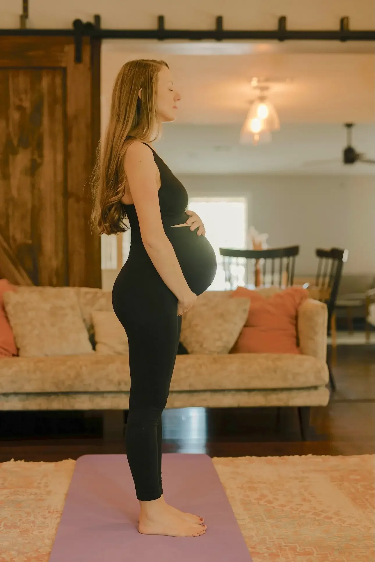 A pregnant woman with long brown hair wearing a black tank top and black leggings is standing on a yoga mat in a cozy living room with a beige couch, wooden sliding door, and dining table in the background. She appears to be meditating or practicing yoga with her eyes closed.