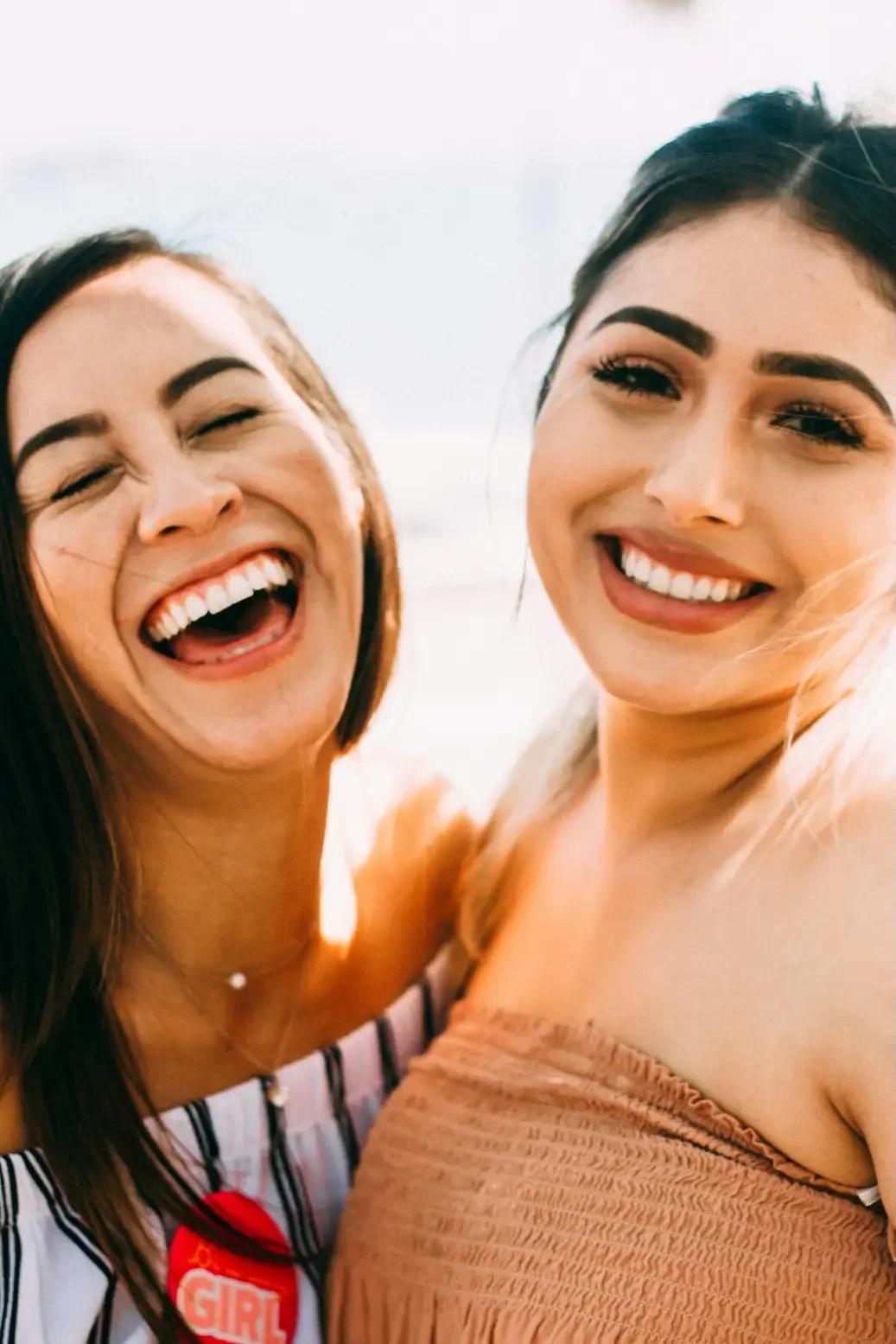 Two women smiling and laughing closely together, with one wearing a striped top and the other in a peach-colored top