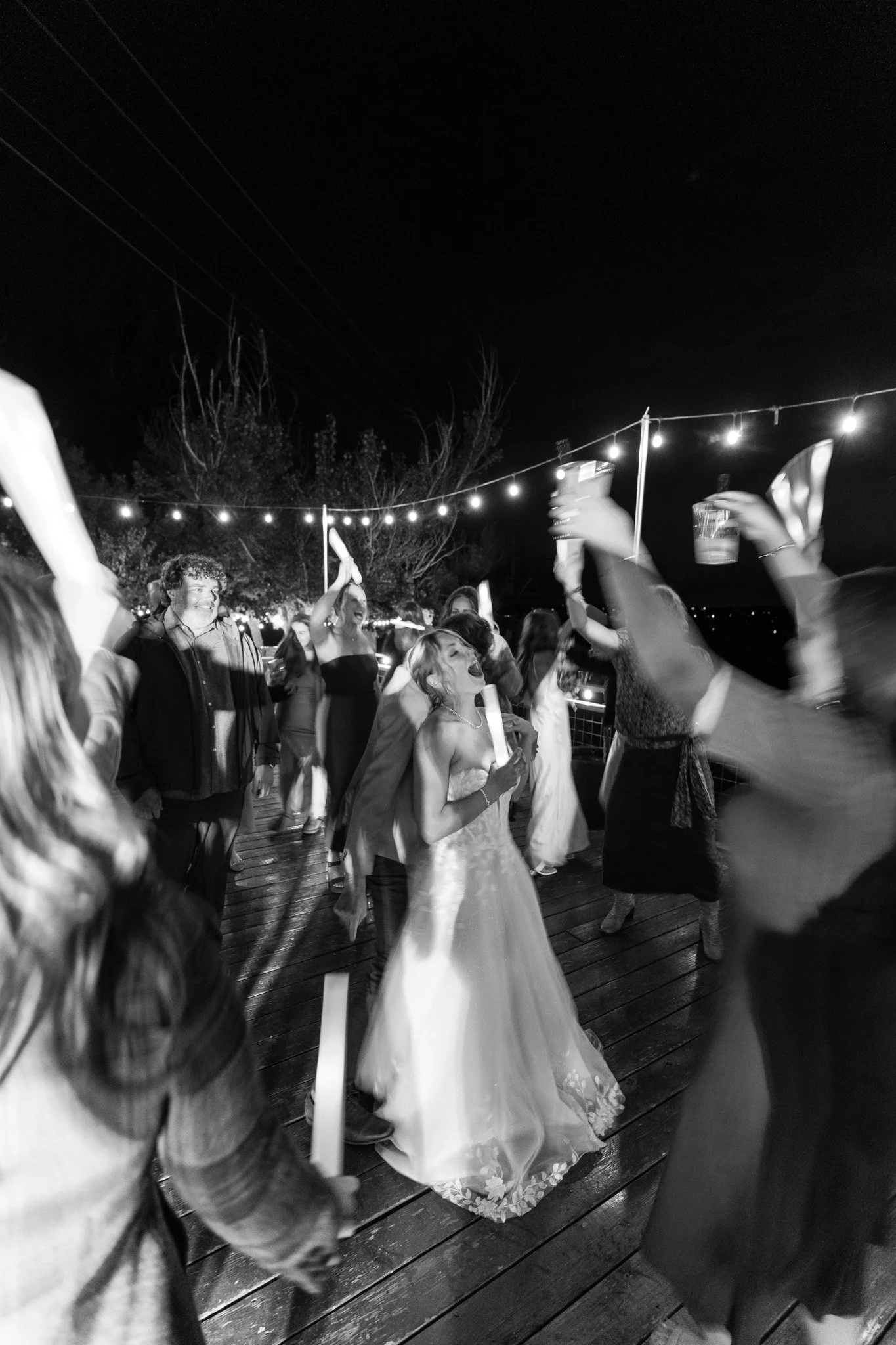 People dancing and celebrating outdoors at night under string lights, with some holding drinks.