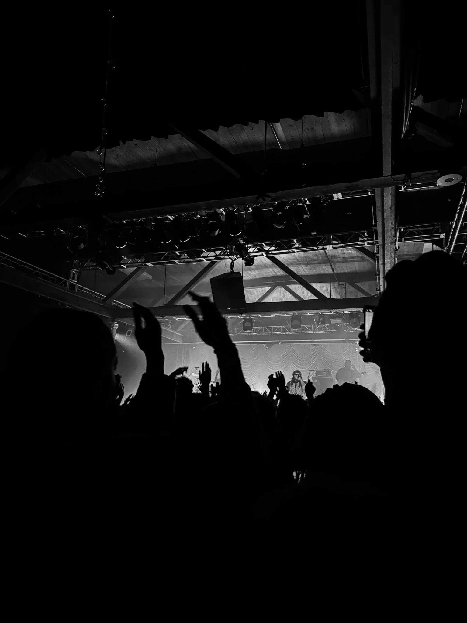 Black and white photo of a concert or event with silhouettes of people with raised hands in the foreground, stage lights, and equipment hanging from the ceiling.