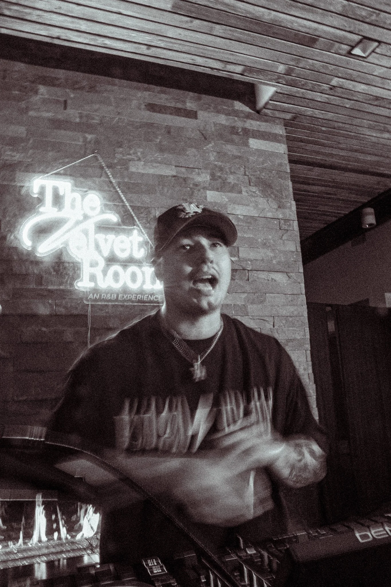 A person playing an electronic keyboard in a dimly lit room with a brick wall and wooden ceiling. There is a neon sign that reads 'The Velvet Room' on the brick wall.