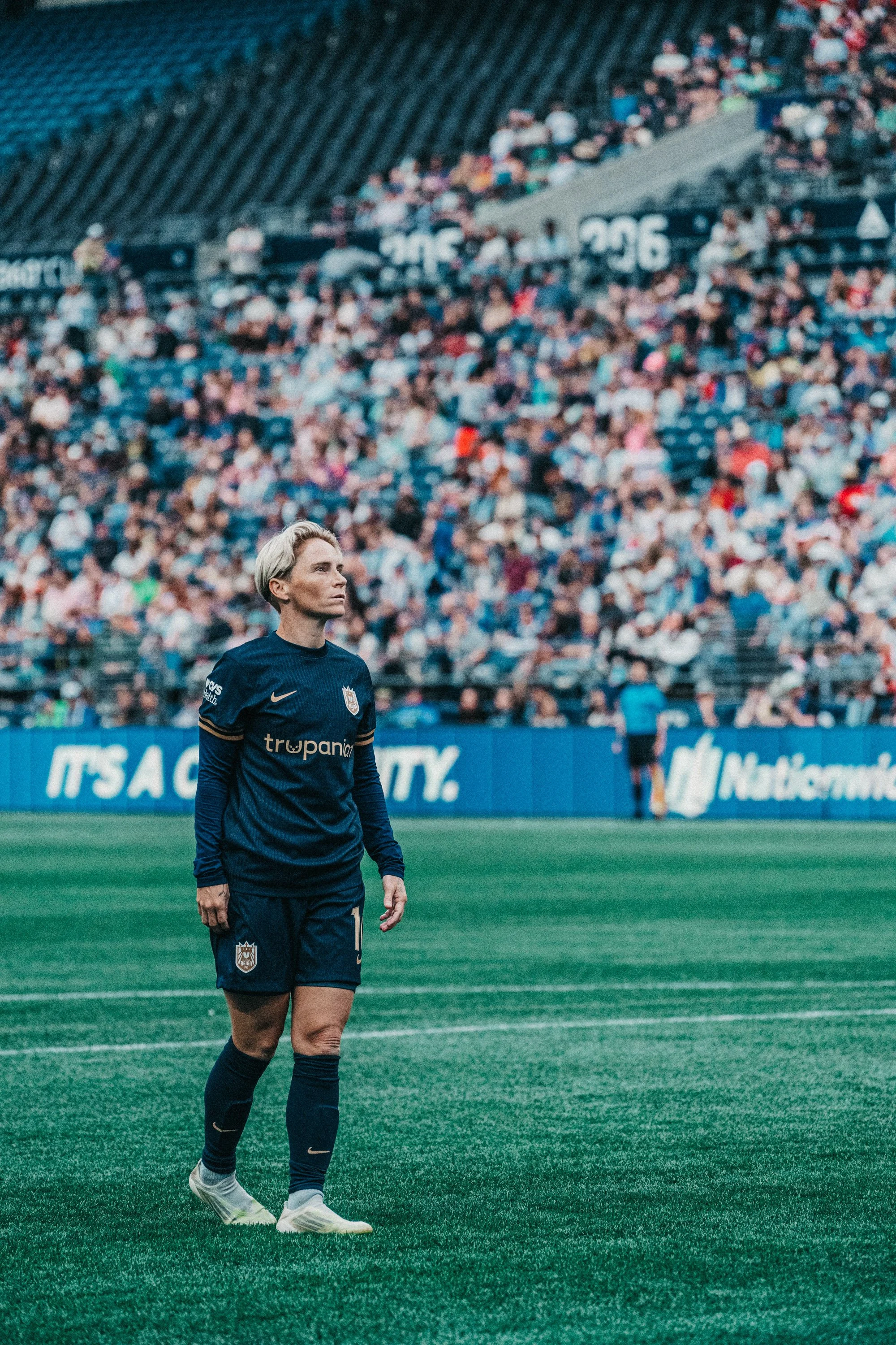 Female soccer player standing on the field during a game, wearing a dark blue uniform with the number 7 and a crest, with a large crowd watching in the background.