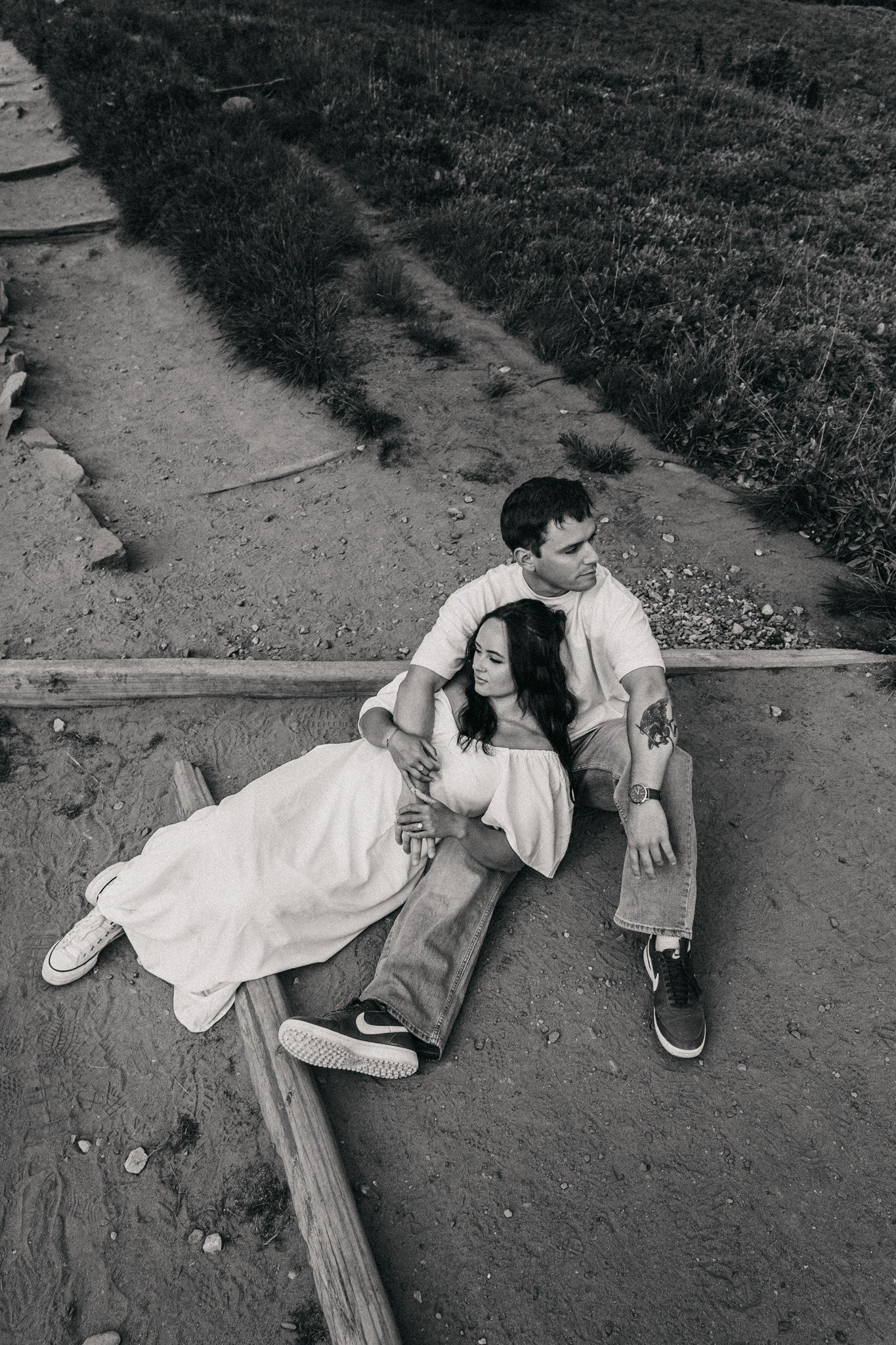 A black and white photo of a man and woman resting on the ground beside a wooden plank, with a grassy and dirt area behind them. The woman lies with her head on the man's lap, and they are embracing each other.