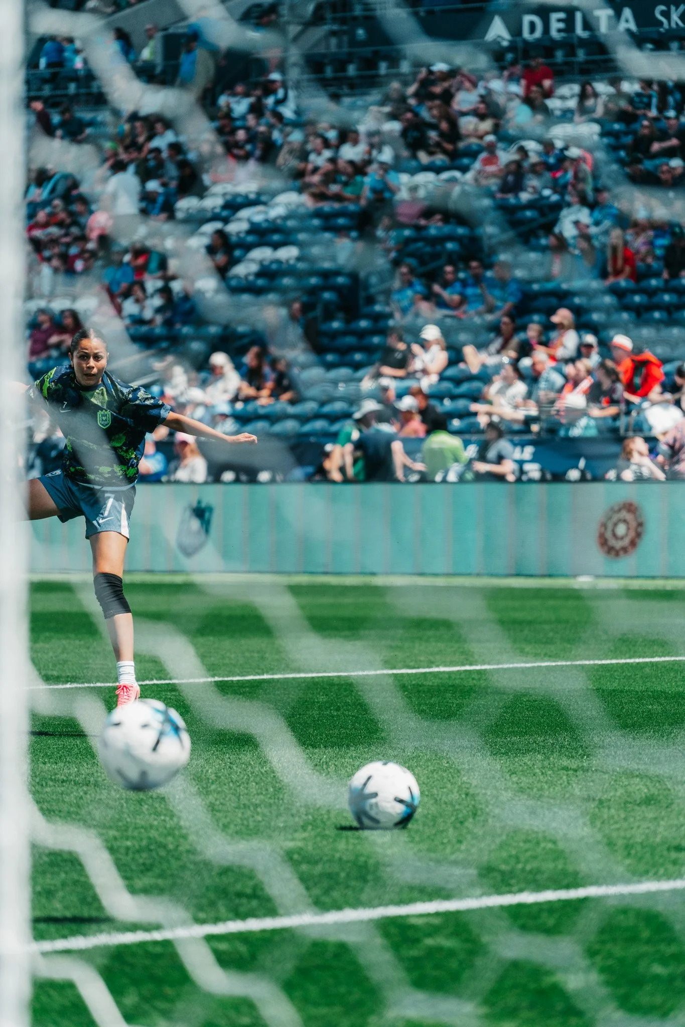 Soccer player kicking soccer balls on the field, with audience in the stands behind the goal.