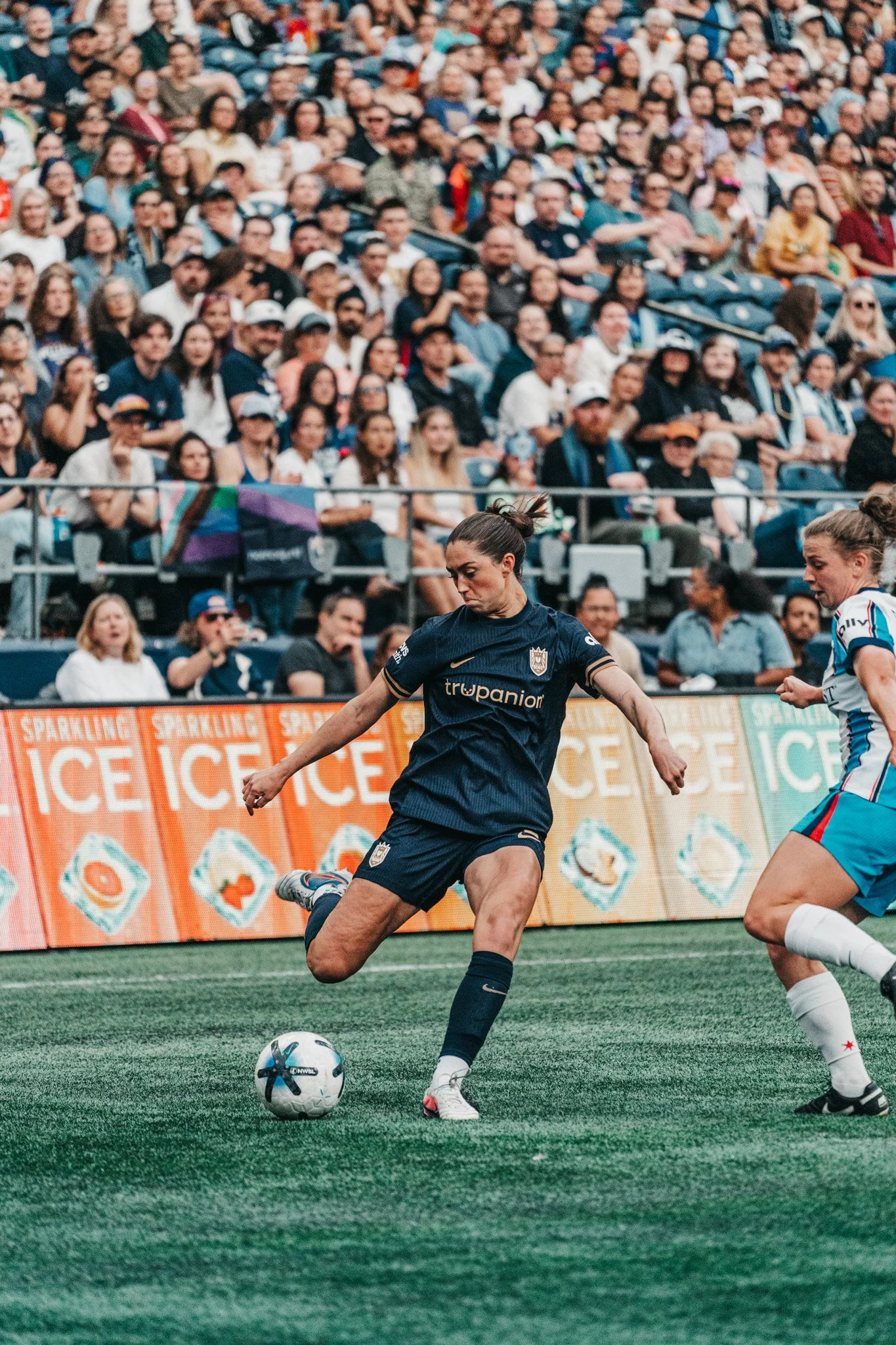 A female soccer player in a navy blue uniform is kicking a soccer ball on a green field, with a crowd of spectators in the background.