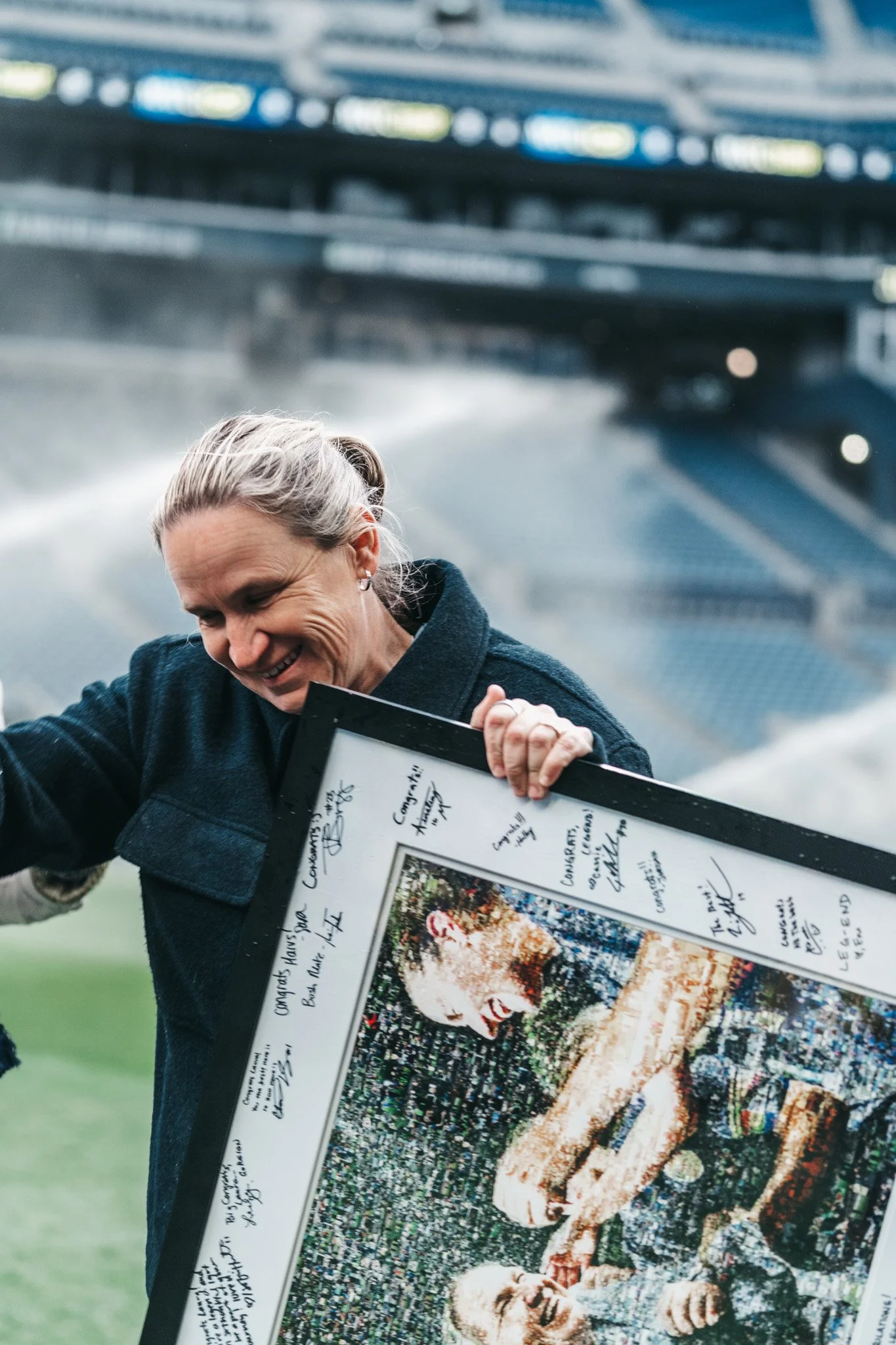 A woman smiling while holding a large, framed poster or certificate with signatures and a colorful image, outdoors on a sports field with a stadium in the background.