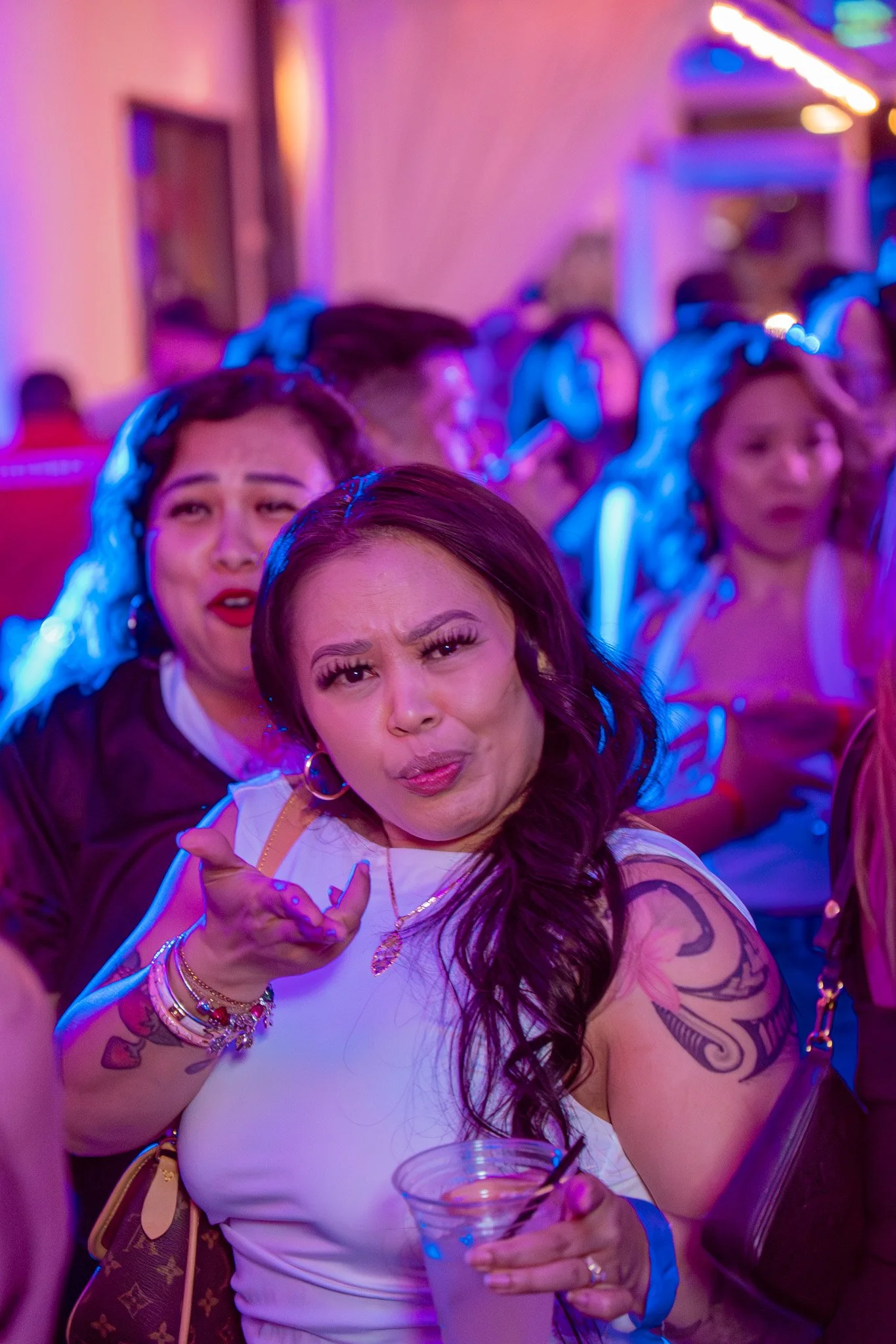 Two women in a crowded bar or party, with purple and blue lighting. One woman in the foreground is making a confused or surprised face, with dark brown hair and tattoos on her arm, holding a clear plastic cup. The woman behind her has dark hair with 