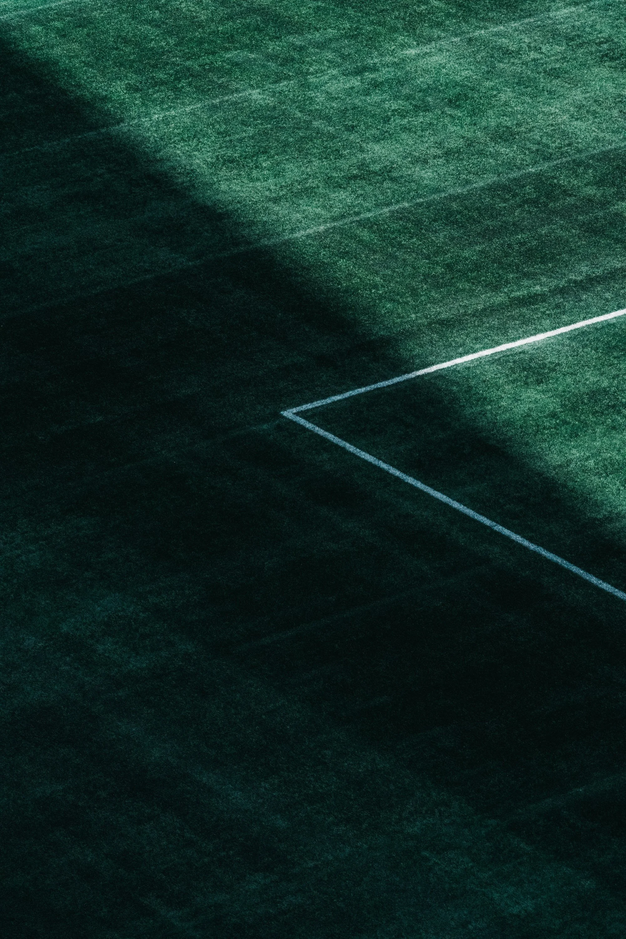 A dark green tennis court with white boundary lines, mostly shadowed with a faint outline of the net.