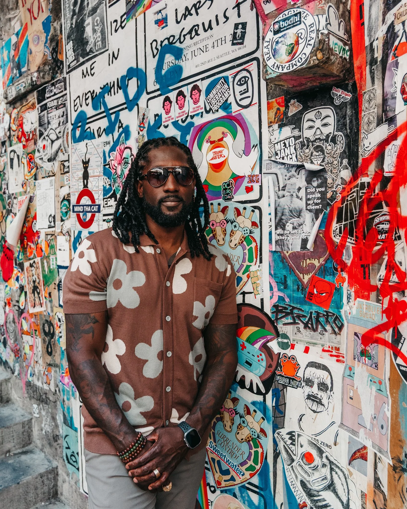 A man with dreadlocks, sunglasses, and a brown and white patterned shirt standing in front of a wall covered with colorful graffiti and stickers.