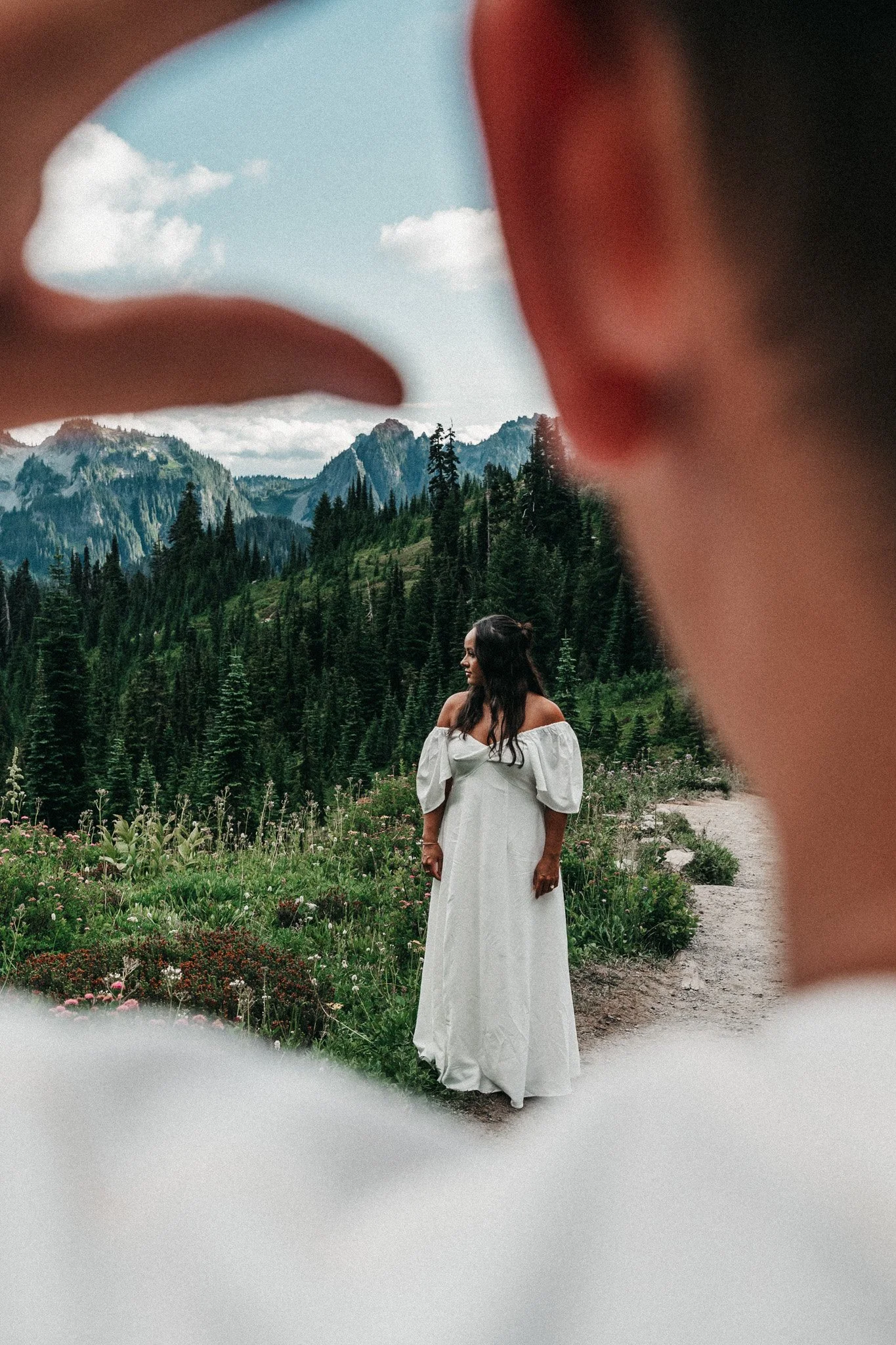 Woman in white dress standing outdoors in front of a mountain landscape with dense green trees and rocky peaks, seen through a frame created by a person's fingers.
