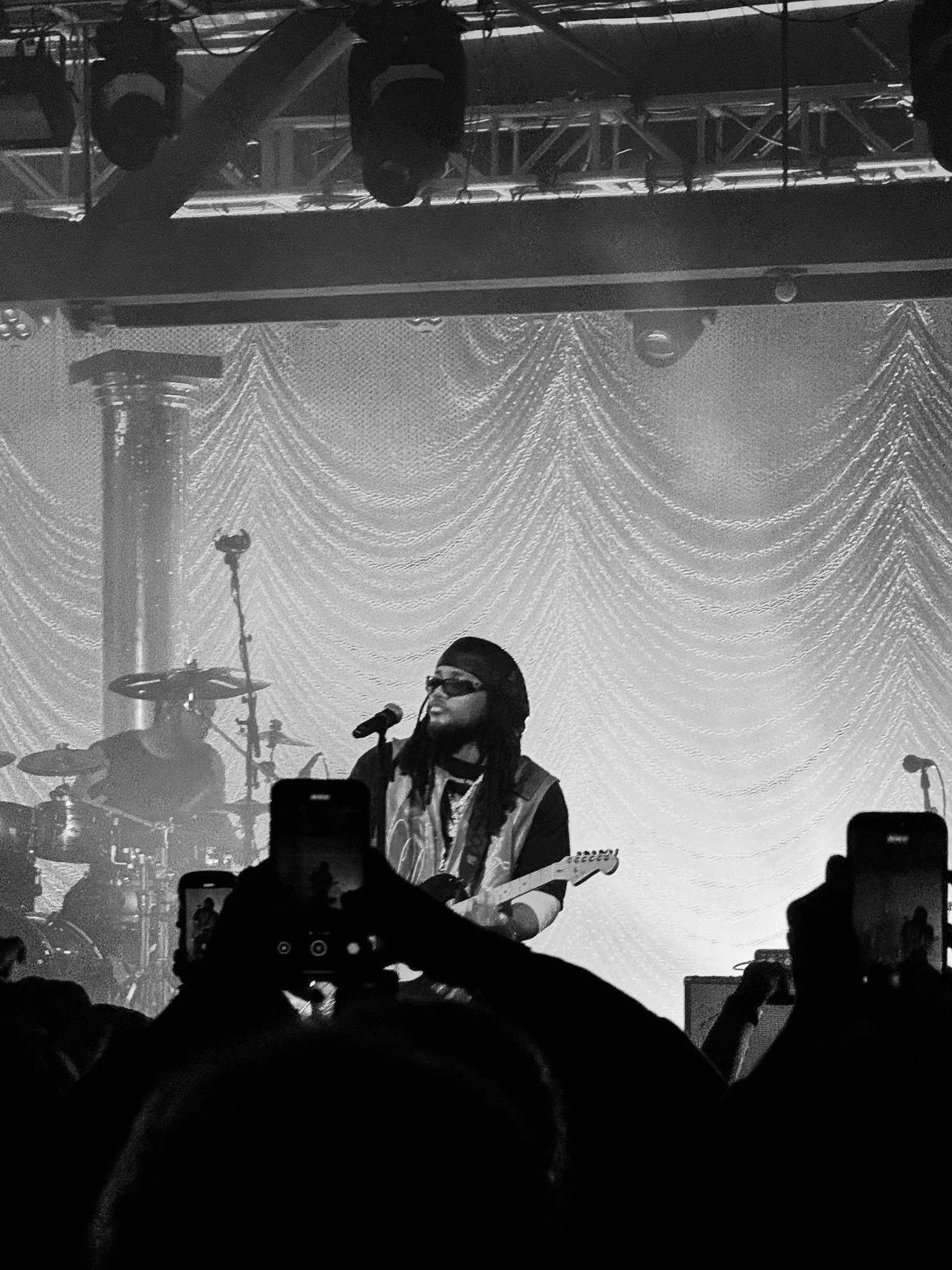 A black and white photo of a female musician playing the guitar and singing into a microphone on stage, with drums in the background and an audience in front taking photos.