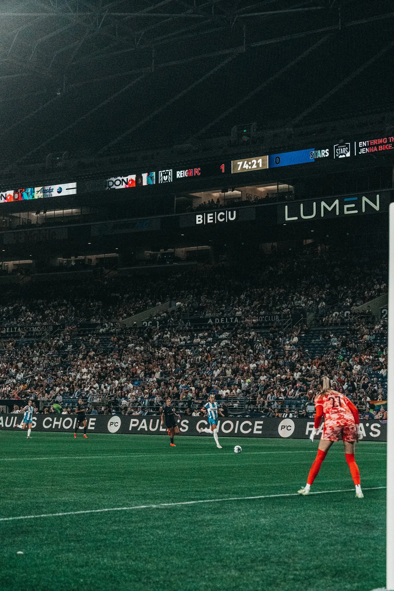Soccer player in red jersey on a green field, while players in blue and white jerseys are in the background, with a large crowd of spectators in the stands and electronic advertising boards displaying various logos and texts.