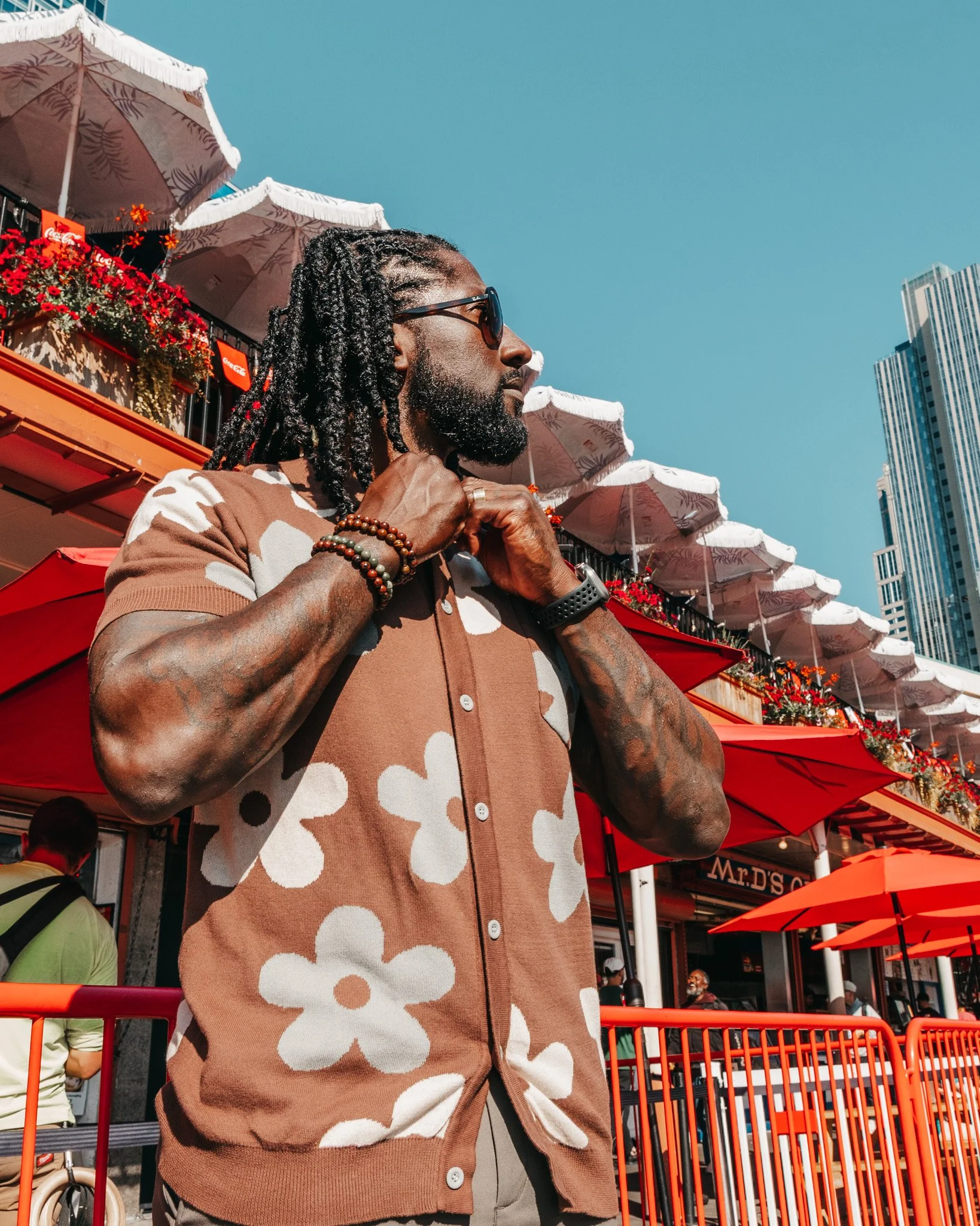 A man with dreadlocks and sunglasses wearing a brown and white floral-patterned cardigan and layered beaded bracelets on his wrist, standing outside near red fencing and umbrellas with a cityscape in the background.