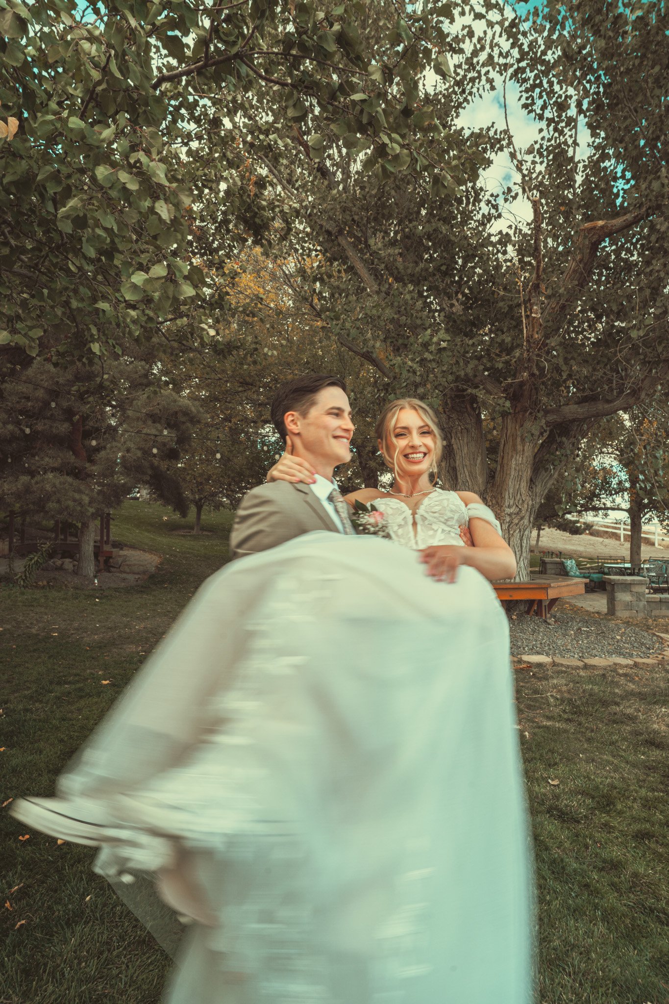 A couple dressed in wedding attire smiling under a large tree in a park setting, with greenery and benches in the background.