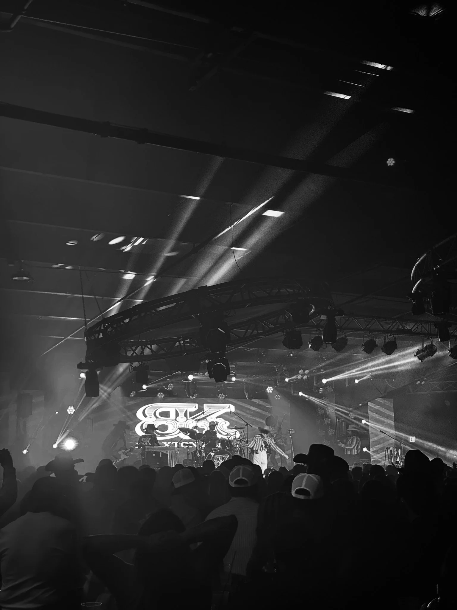 Stage with bright lights, a large screen displaying a logo, and musical performers with instruments in front of an audience wearing caps.