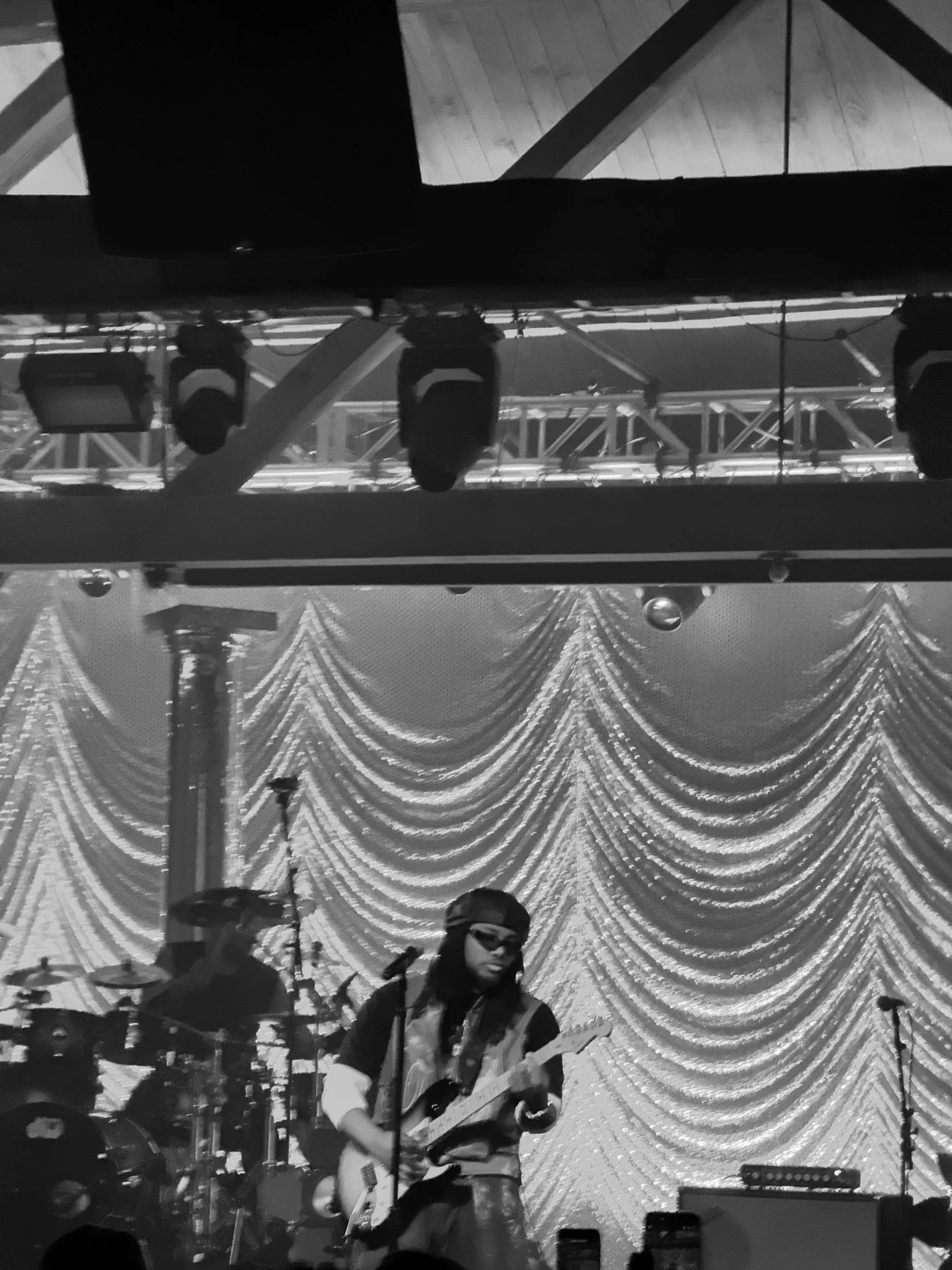 A person playing an electric guitar on stage, with a microphone in front, set against a backdrop of draped, shimmering curtains and stage lighting.