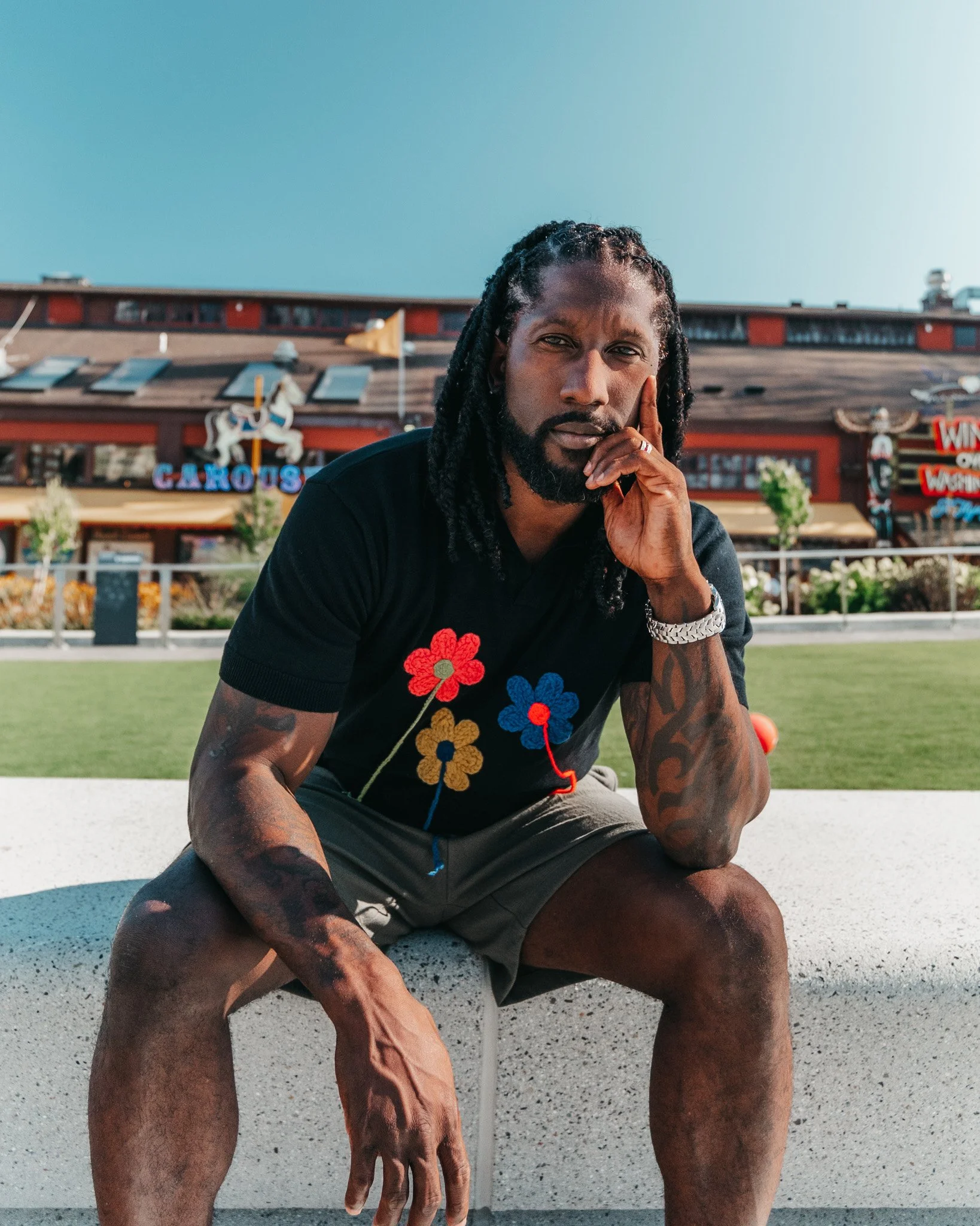A man with long dreadlocks and a beard sits outside on a concrete surface, resting his chin on his hand and looking at the camera with a serious expression. He wears a black T-shirt with colorful flower graphics and a silver watch. In the background,