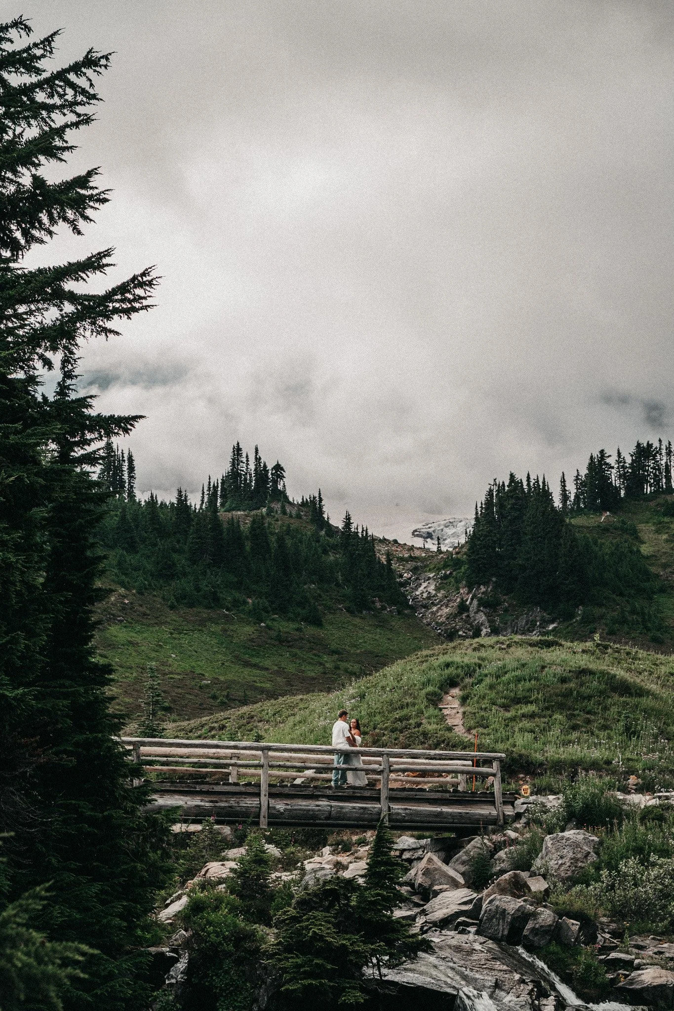 A couple stands on a wooden bridge over a rocky stream in a lush green forest, with mountains and cloudy sky in the background.