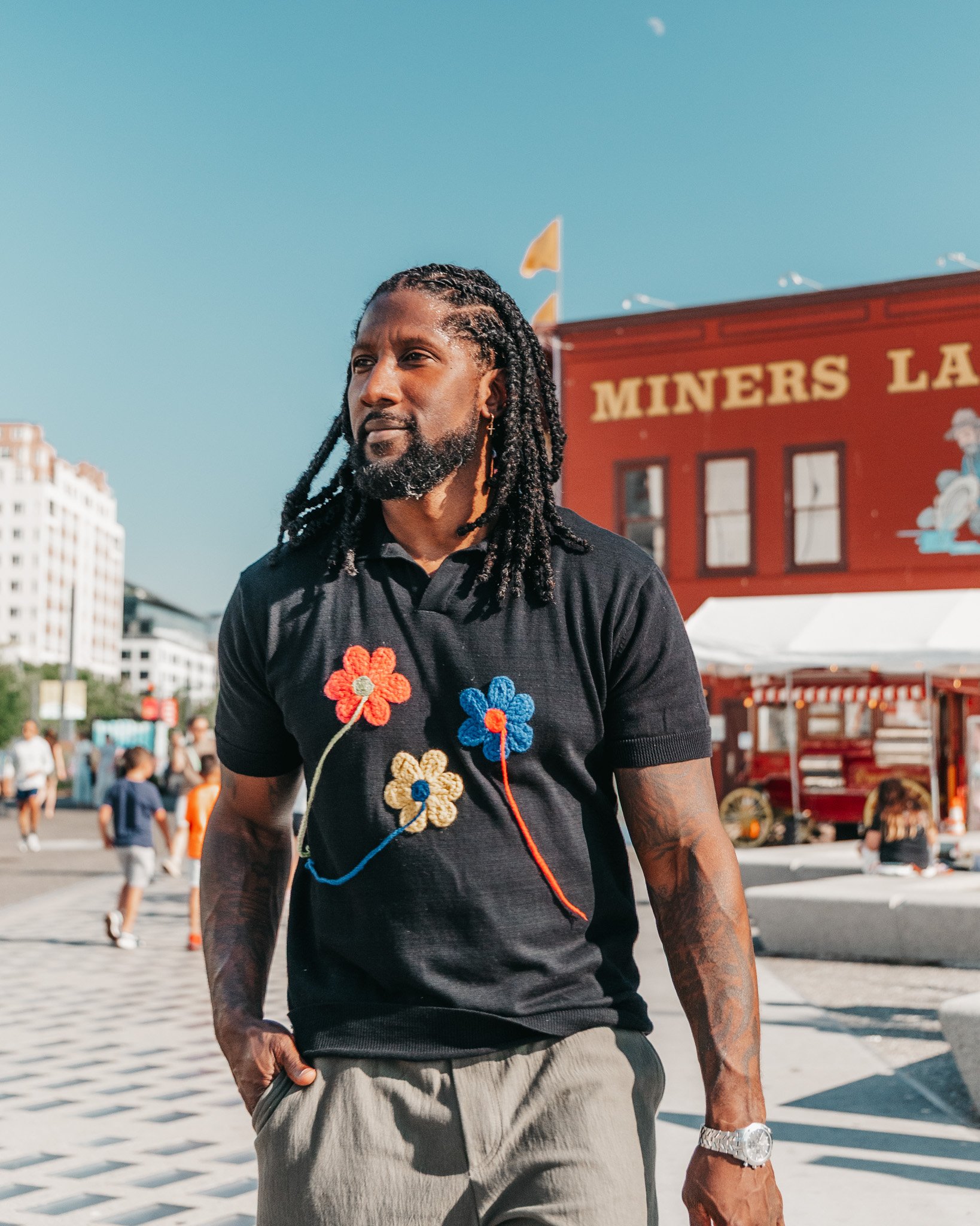 A man with long dreadlocks, a beard, and earrings standing in front of a red building with the sign 'Miners' at an outdoor event. People are in the background. The man is wearing a black shirt with colorful crochet flowers, a watch, and has his left 