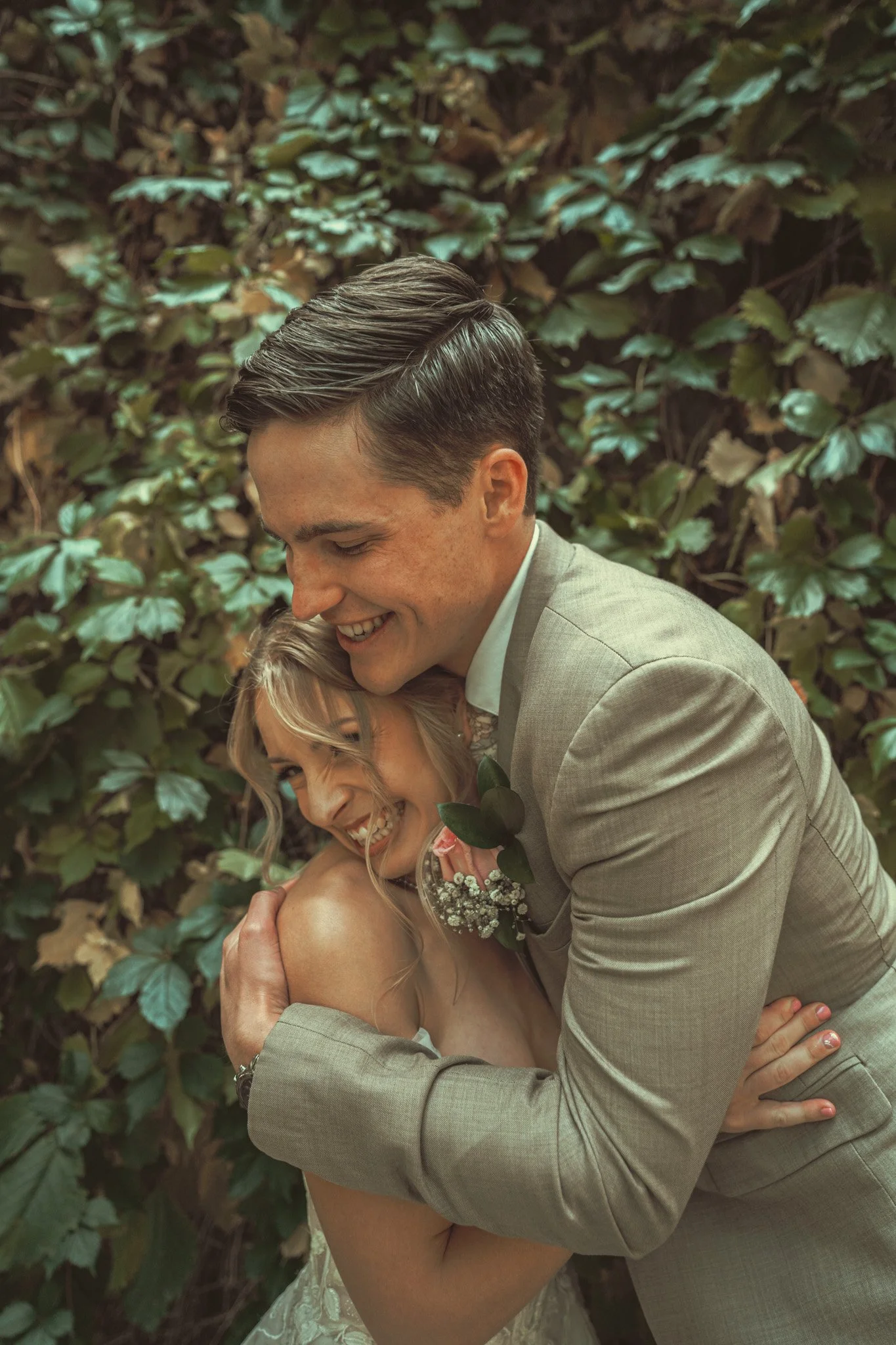 A smiling man in a gray suit embracing a blonde woman with curly hair, who is holding a small bouquet of flowers, against a background of green leafy foliage.