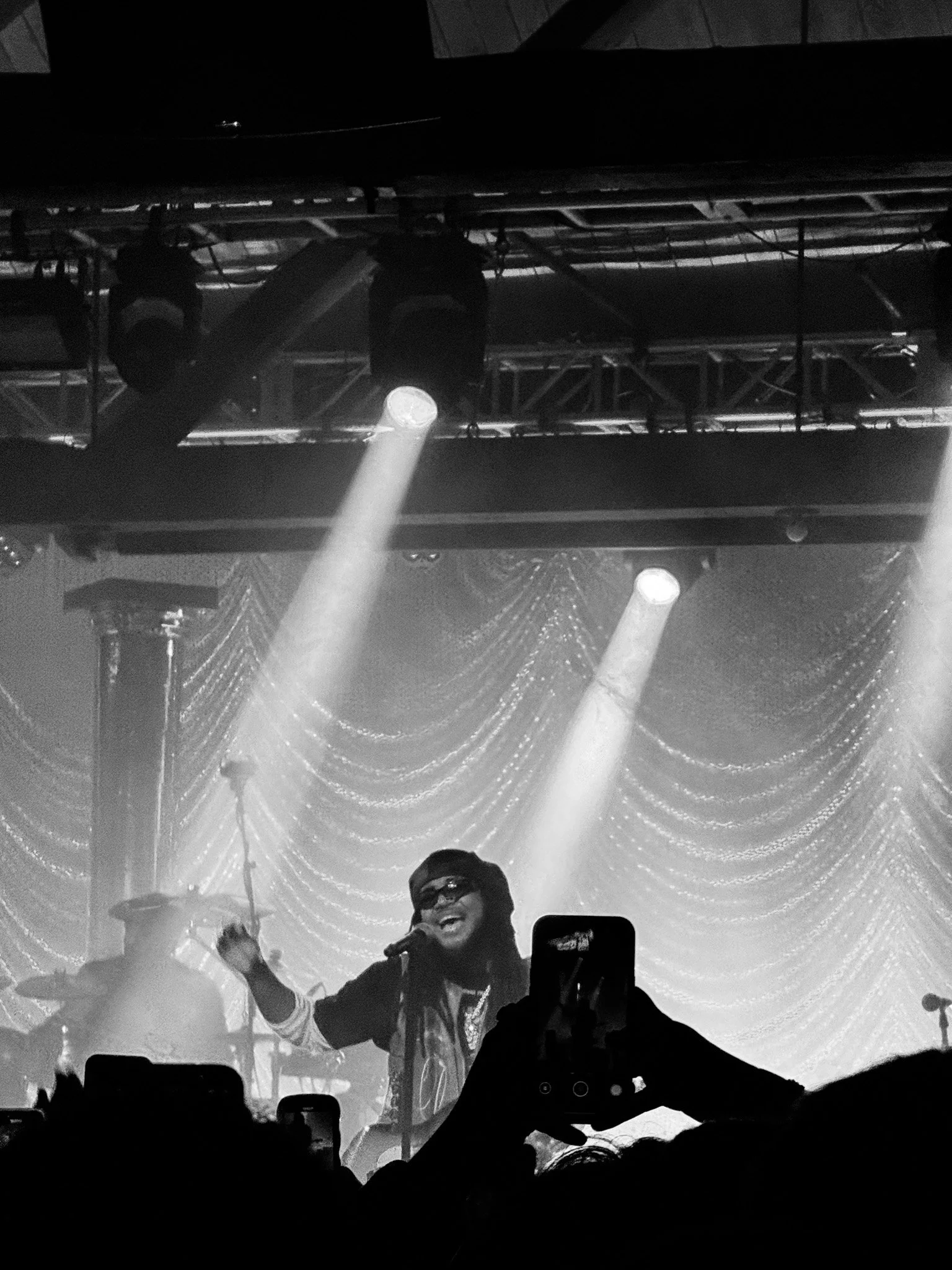 A performer on stage holding a microphone, smiling, with colorful lighting and decorative curtains in the background.