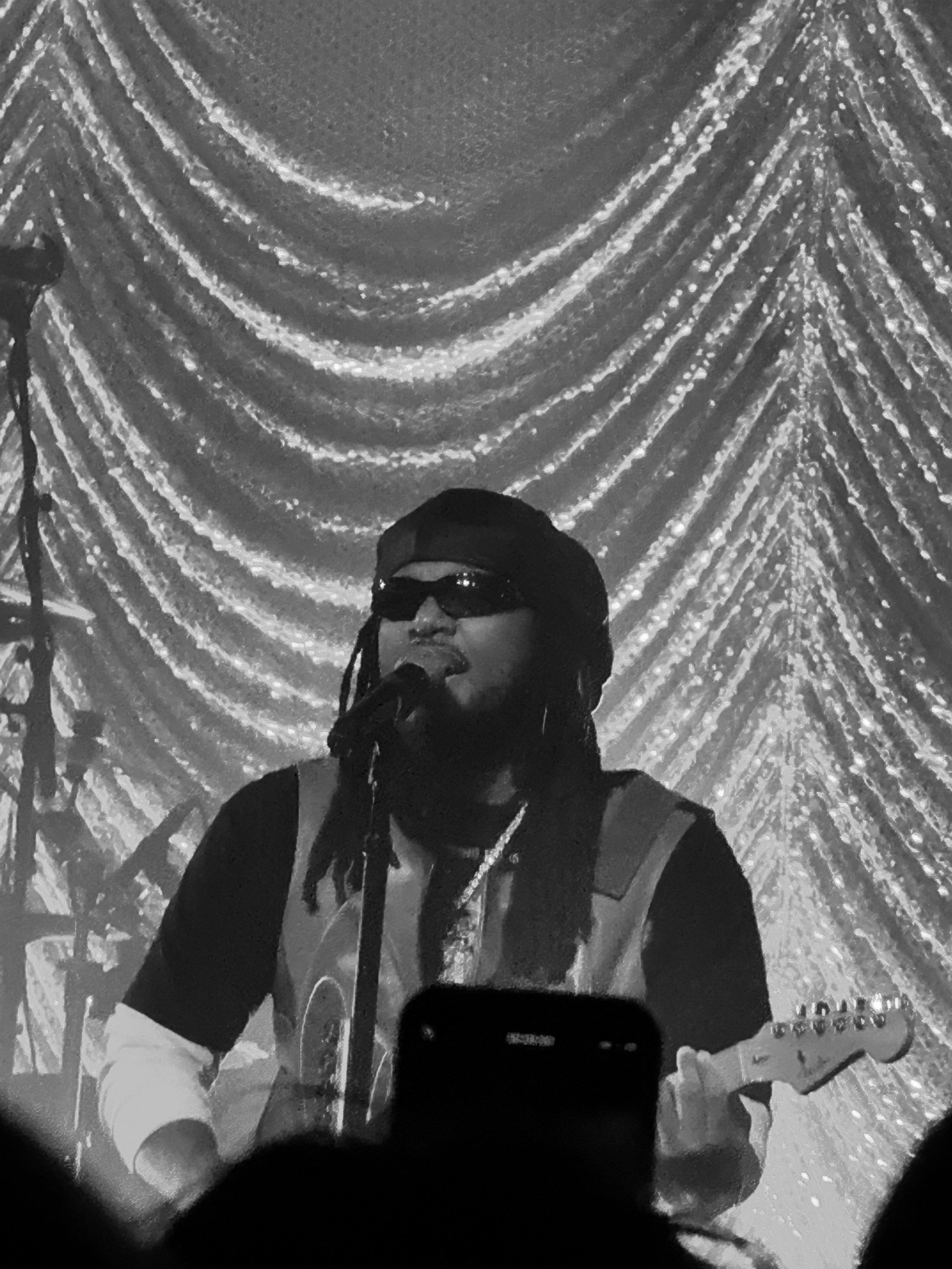 A person wearing sunglasses, a cap, and a winter jacket, taking a selfie in front of a shimmering, possibly metallic or lights-decorated backdrop.