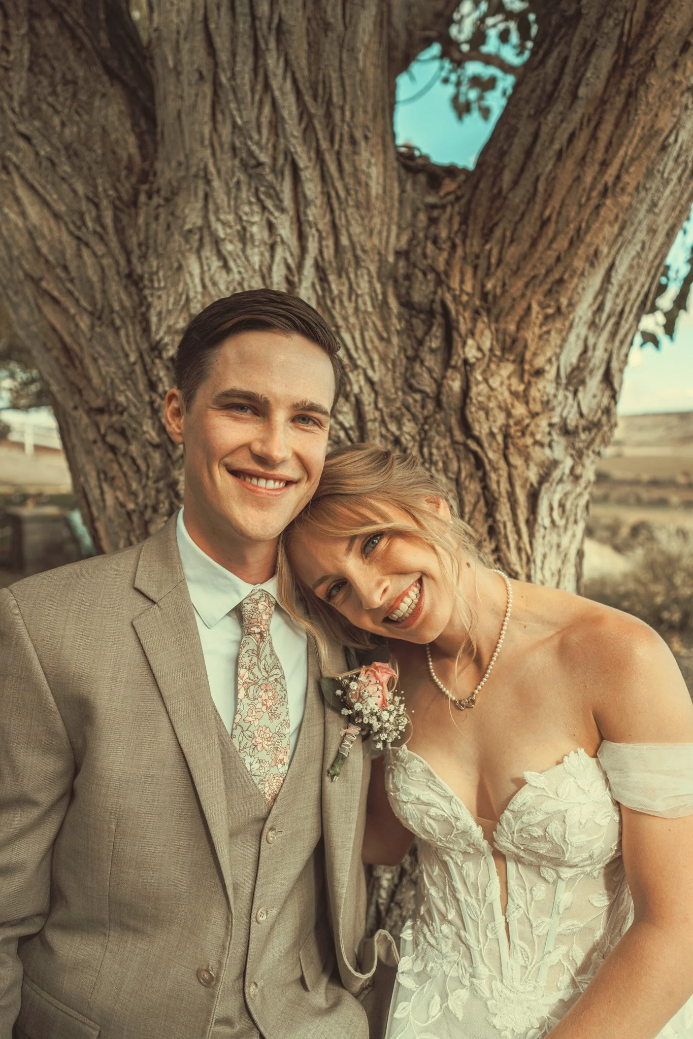 A smiling couple, the man in a beige suit with a floral tie, and the woman in a white wedding dress with lace details, are standing close together under a large tree with textured bark outdoors.