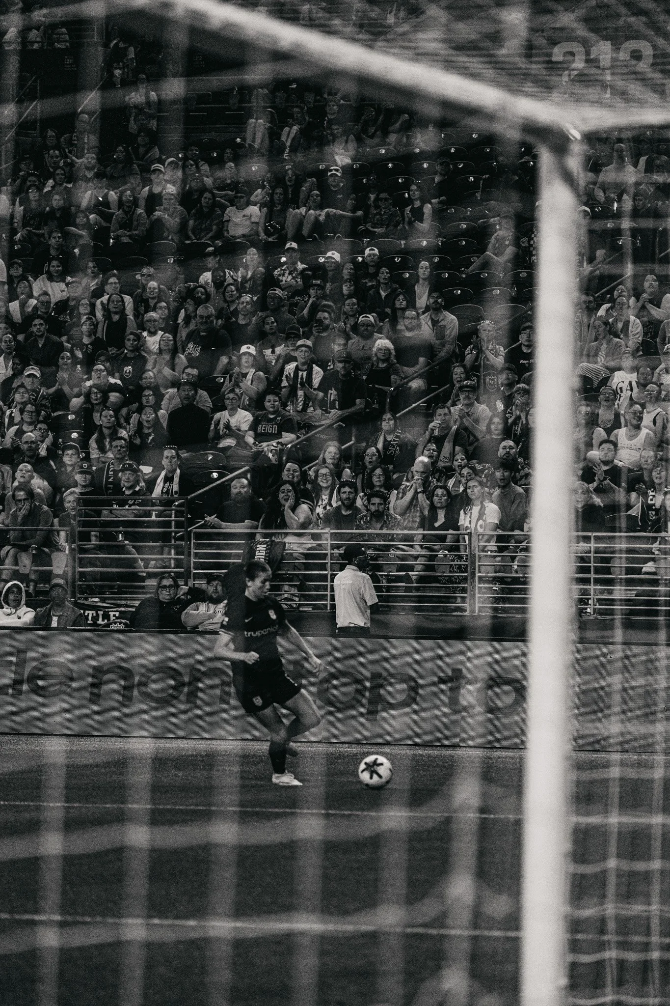 A black and white photo of a soccer game, showing a player near the goal with a ball, and a stadium filled with spectators watching the game.