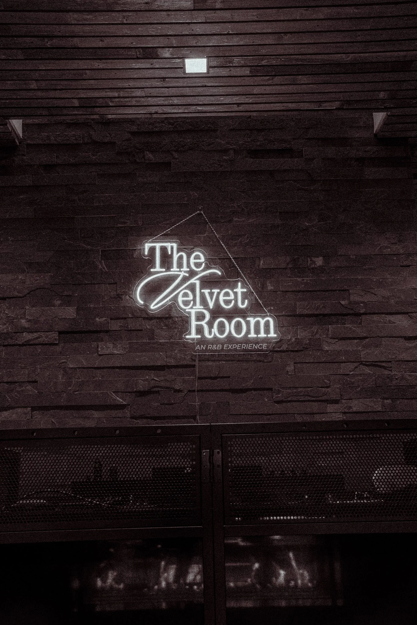 A neon sign on a dark brick wall that reads 'The Velvet Room' with smaller text underneath that says 'An R&B Experience'.